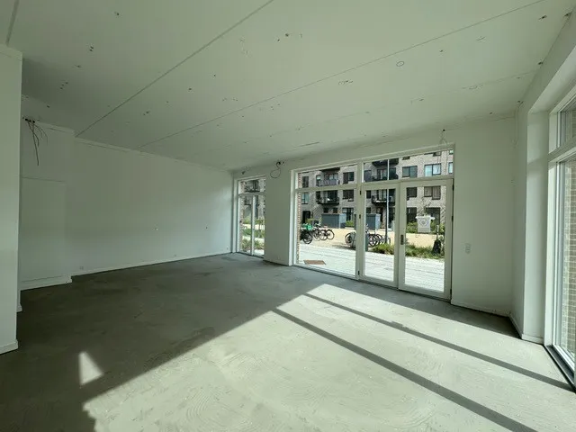 En tom stue med hvide vægge og gulv samt store glasruder til gaden