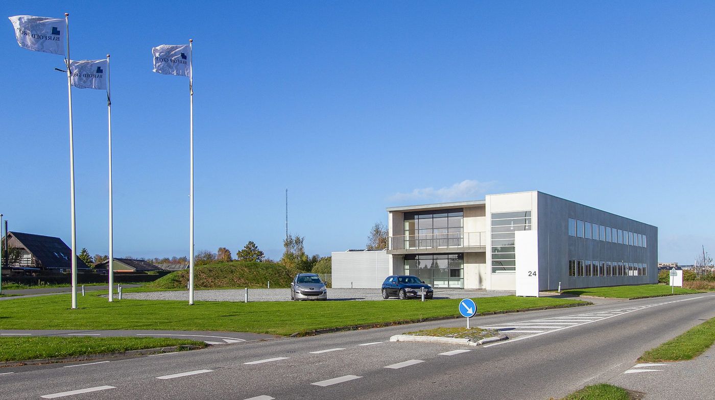 To biler parkeret ved et modernt og hvidt bygningskompleks med tre flag med logoet "Harboe Bryggerierne"