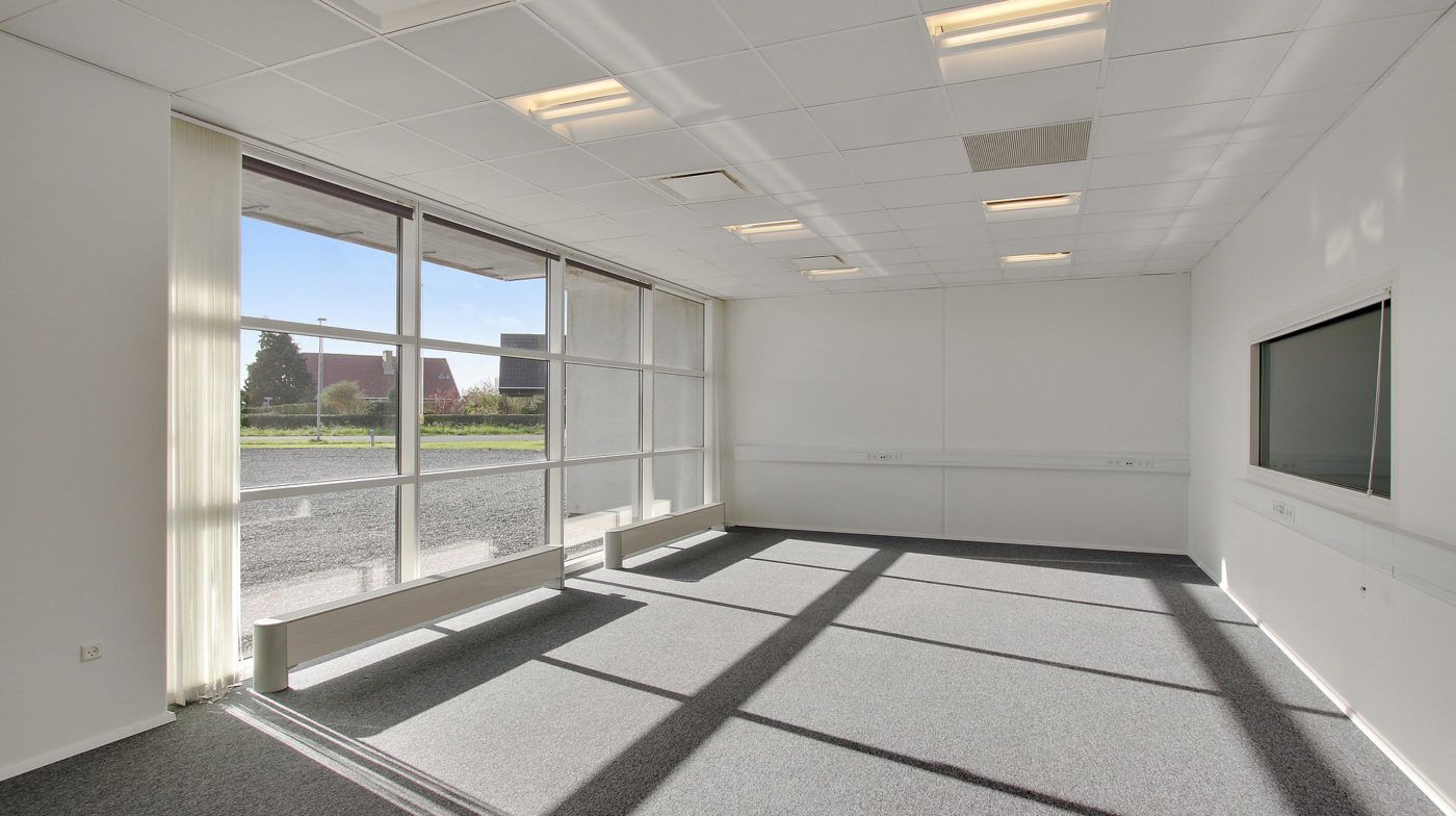 Store hvide vægge og loft i en lukket stue. Store vinduer med hvide rammer og skyggekast på gulvet. To vægmonterede elstik.