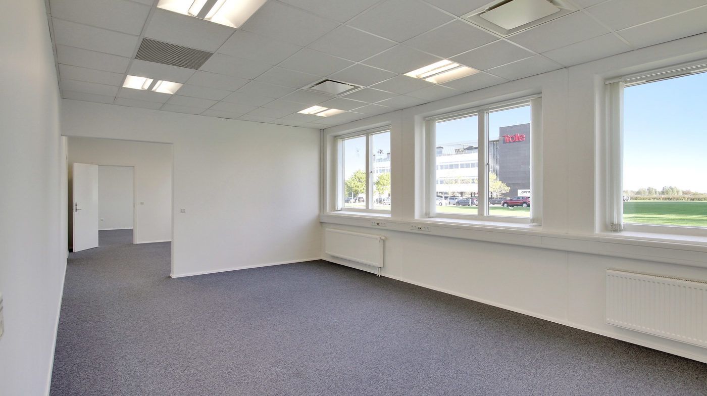 Et stort kontor med hvide vægge og gulv, hvor vinduerne er åbne og udsigten til en park med grønne træer og bygninger