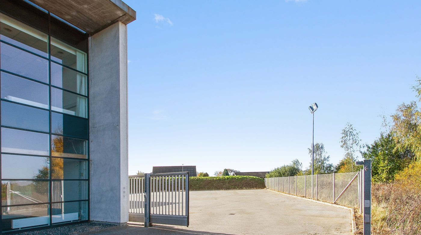 En åben parkeringsplads med glasvinduer og port samt grønne buske og træer bagved