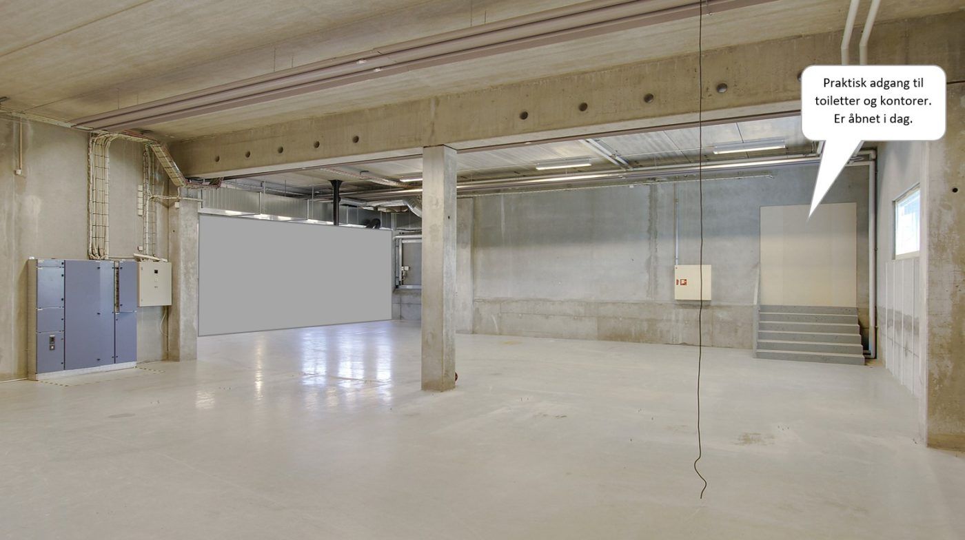 En stor og mørk garage med en hvid væg, en høj søjle og en åbning i loftet, der fører til toiletter og kontor.