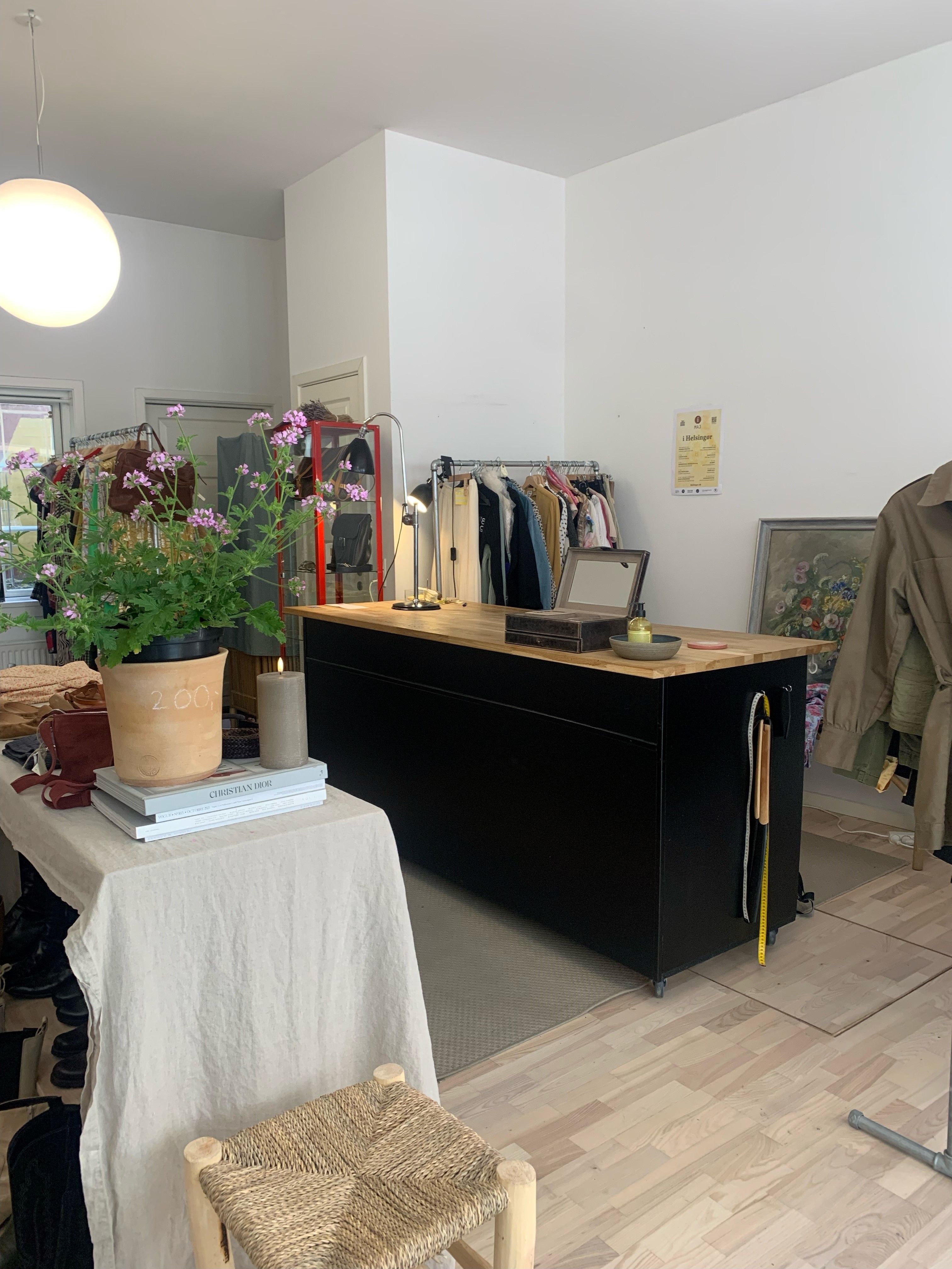 En møbleret butik med en stol, bord og plante med lyserøde blomster samt tøj og sko på hylder og gulvet