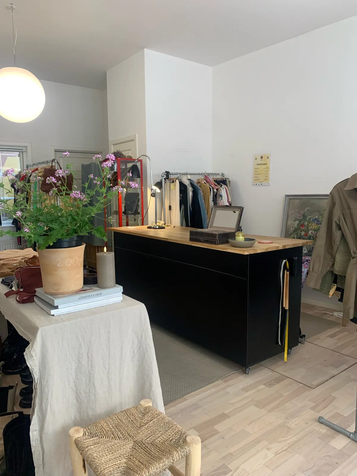 En møbleret butik med en stol, bord og plante med lyserøde blomster samt tøj og sko på hylder og gulvet