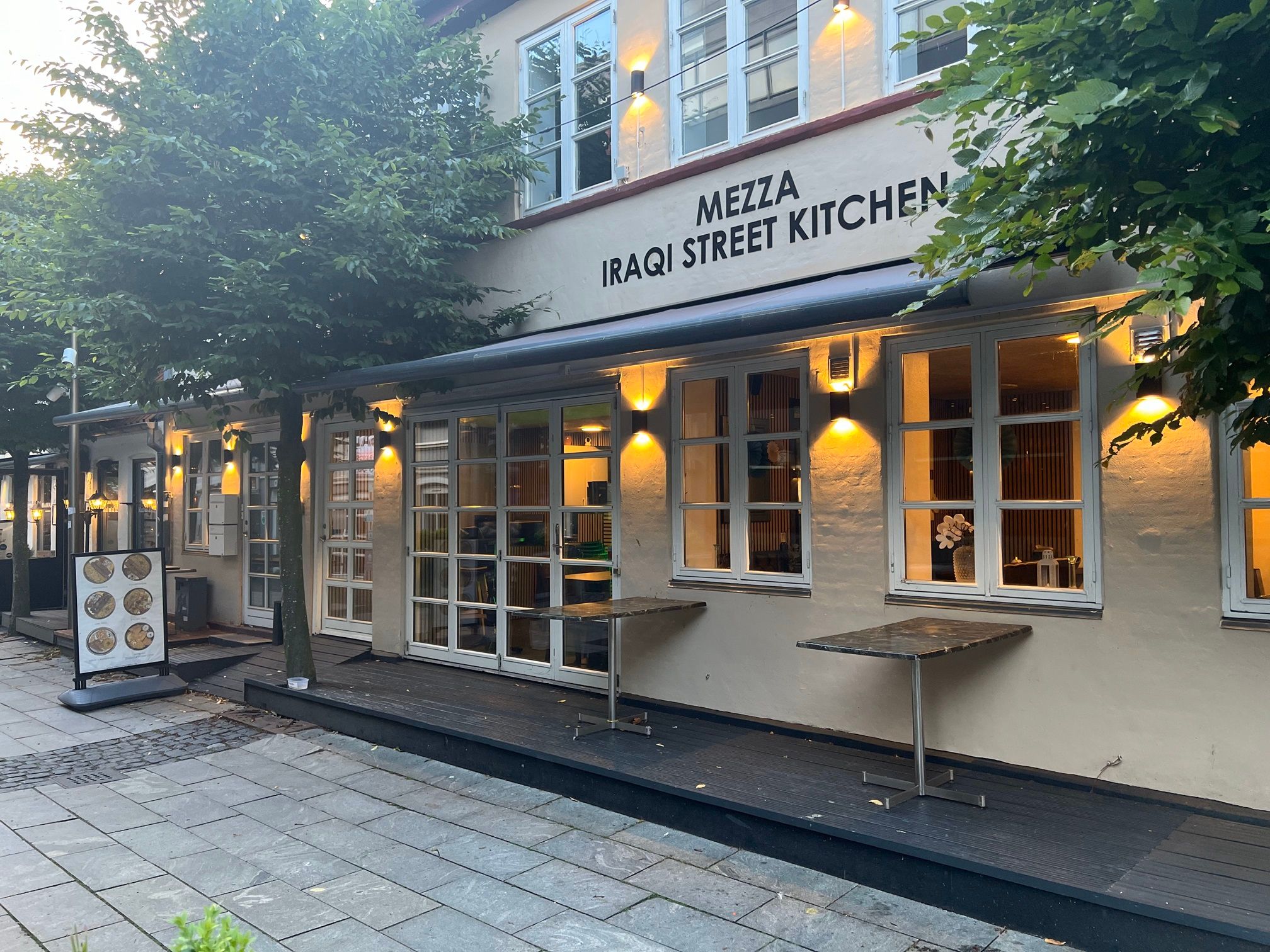 Mezza Iraqi Street Kitchen i Horsens med træer og bord på foran. Det er en restaurant med vinduer og lysekroner.