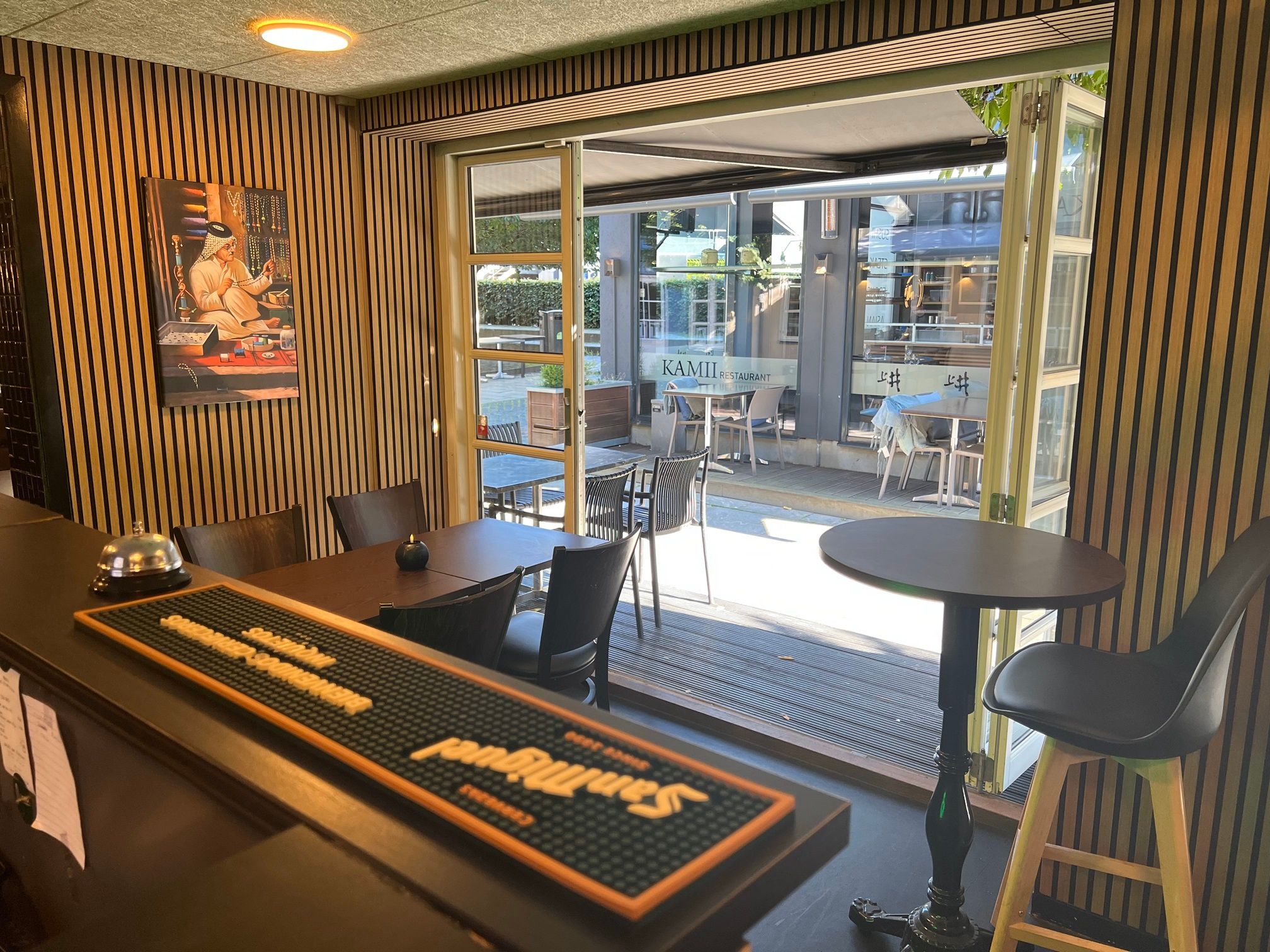 Restaurant med udgang til terrasse med bord og stole samt maleri med guldmynter i en niche