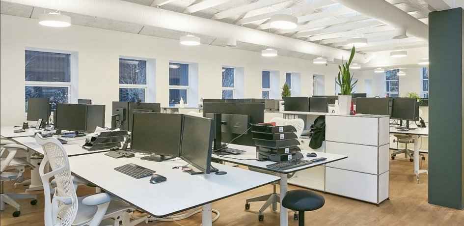 Et moderne kontor med hvide skriveborde og stole, højt op til loftet og lyse højloftede lofter med lysekroner og plante i