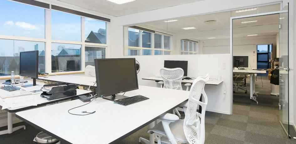 Kontor med moderne indretning og teknologi, med skriveborde, computere og store vinduer, der giver god udsigt.