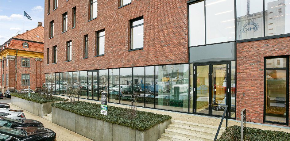 En moderne, rødbrikket bygning med glasdøre og vinduer, foran er et parkeringsområde med flere biler, og der er grønne anlæg