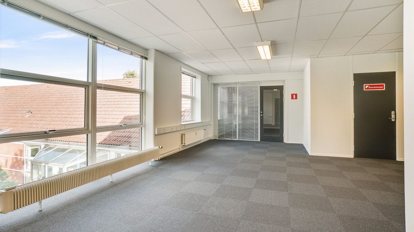 Et tomt kontorlokale med karrevinduer og et radiatorophæng. En lukket dør med en brandalarm. Lyskroner og hvidt loft.