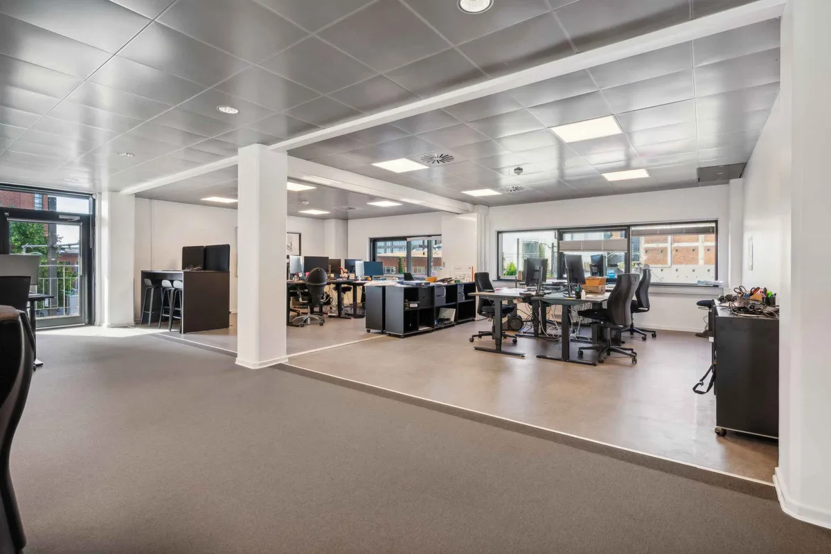 Kontor med højloft, moderne møbler og vinduer med udsigt til byen. Slanke skriveborde, lyse loftslampe og gulvbelægning med