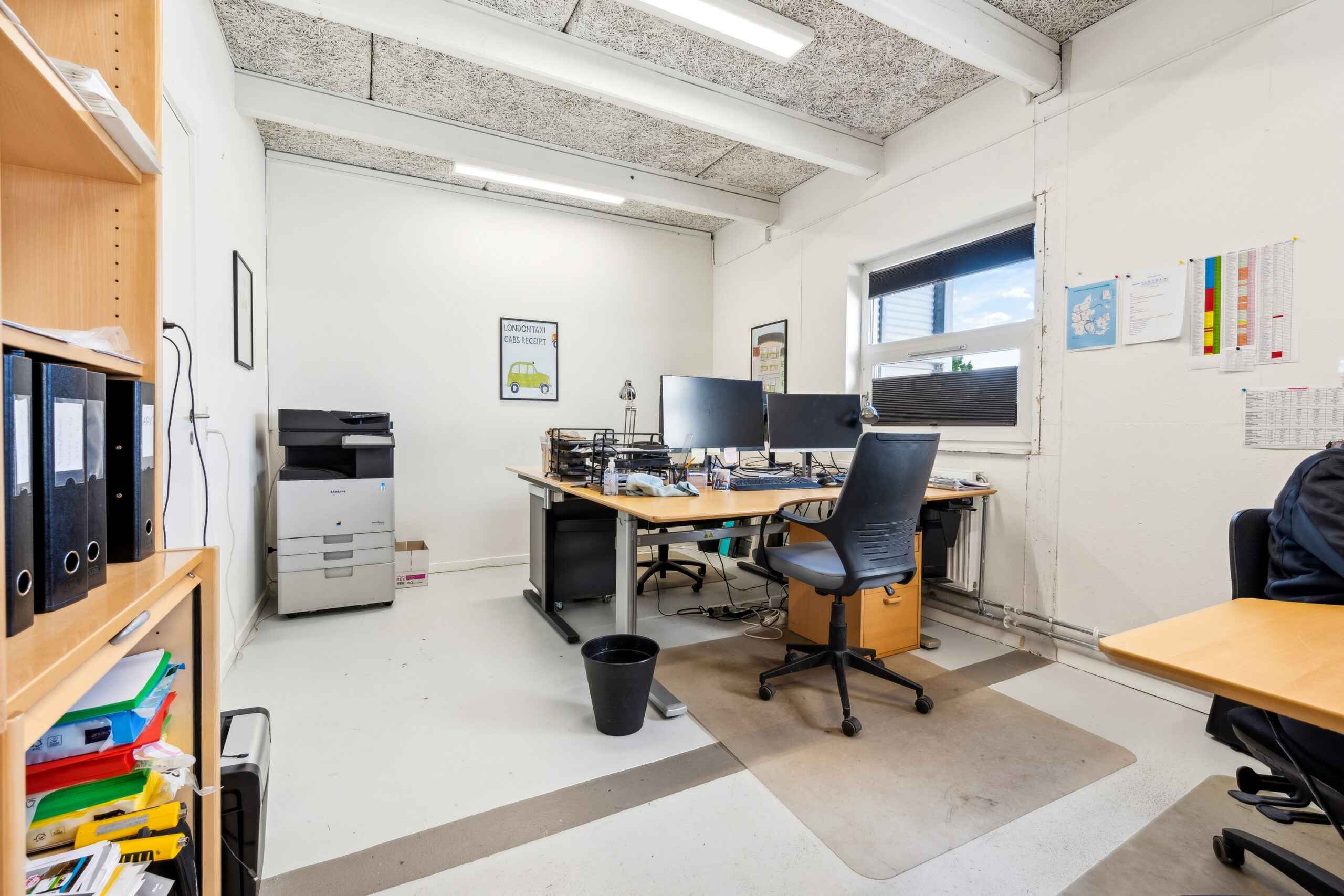 Kontor med skrivebord og computere, højtliggende loftsbælg og lysekroner, vinduer og papirkurv samt skraldespand