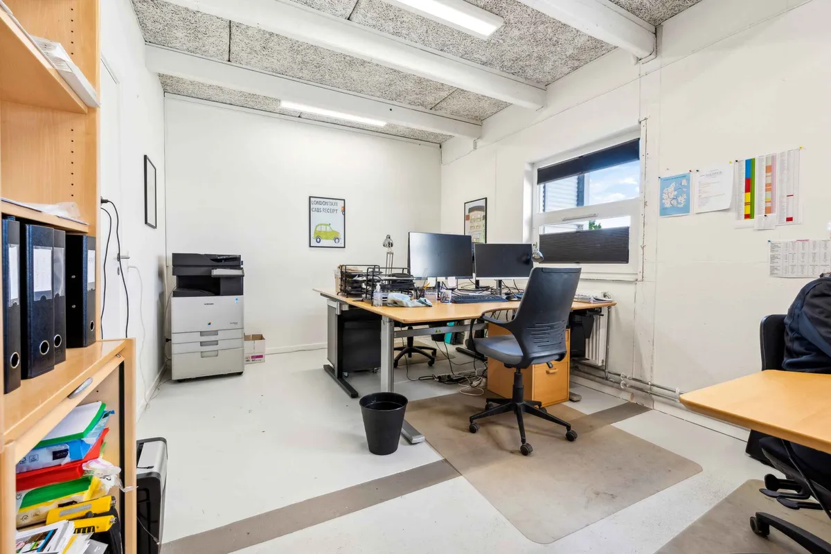 Kontor med skrivebord og computere, højtliggende loftsbælg og lysekroner, vinduer og papirkurv samt skraldespand