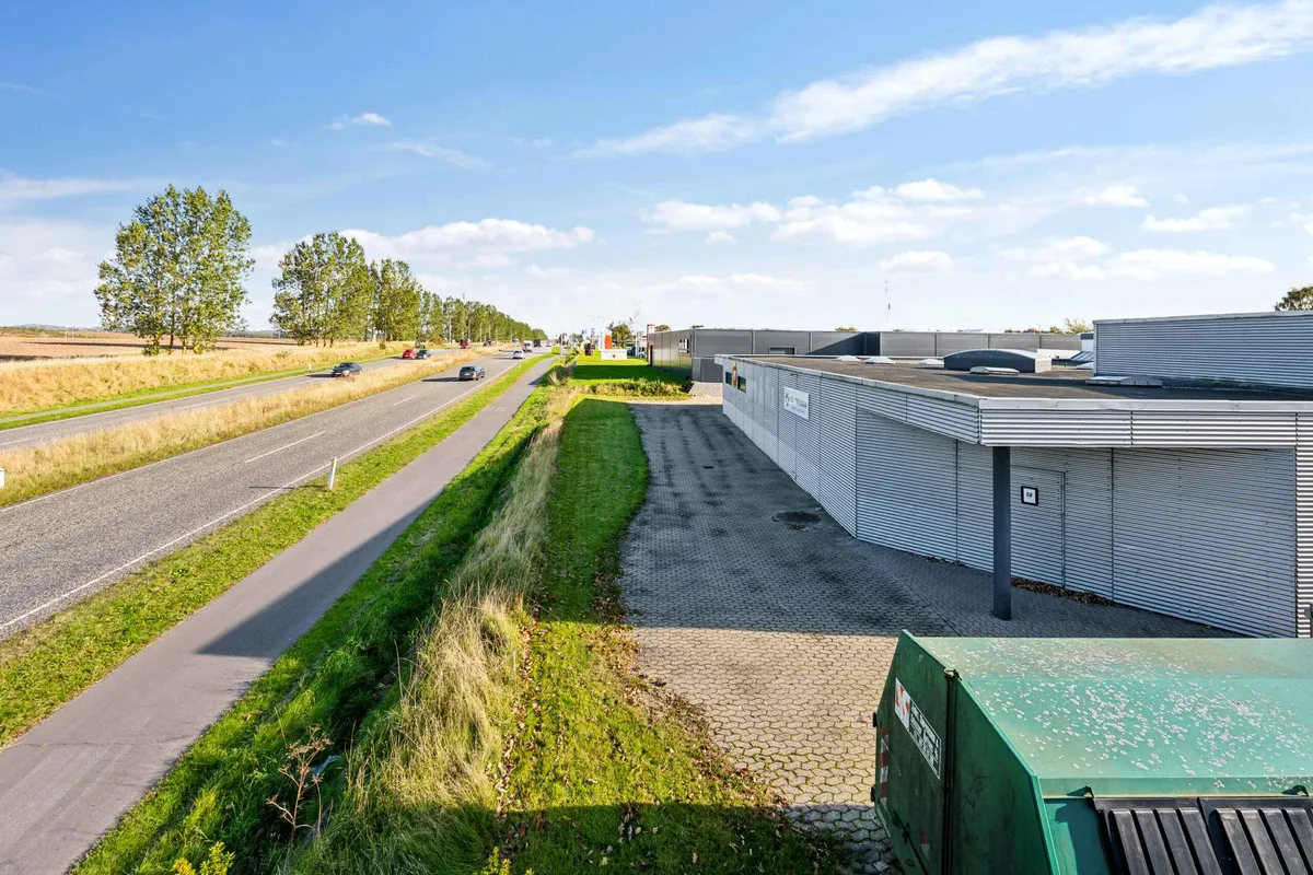 En trafiktæt vej med biler og tog langs siden og en fabriksbygning med parkeringsplads foran