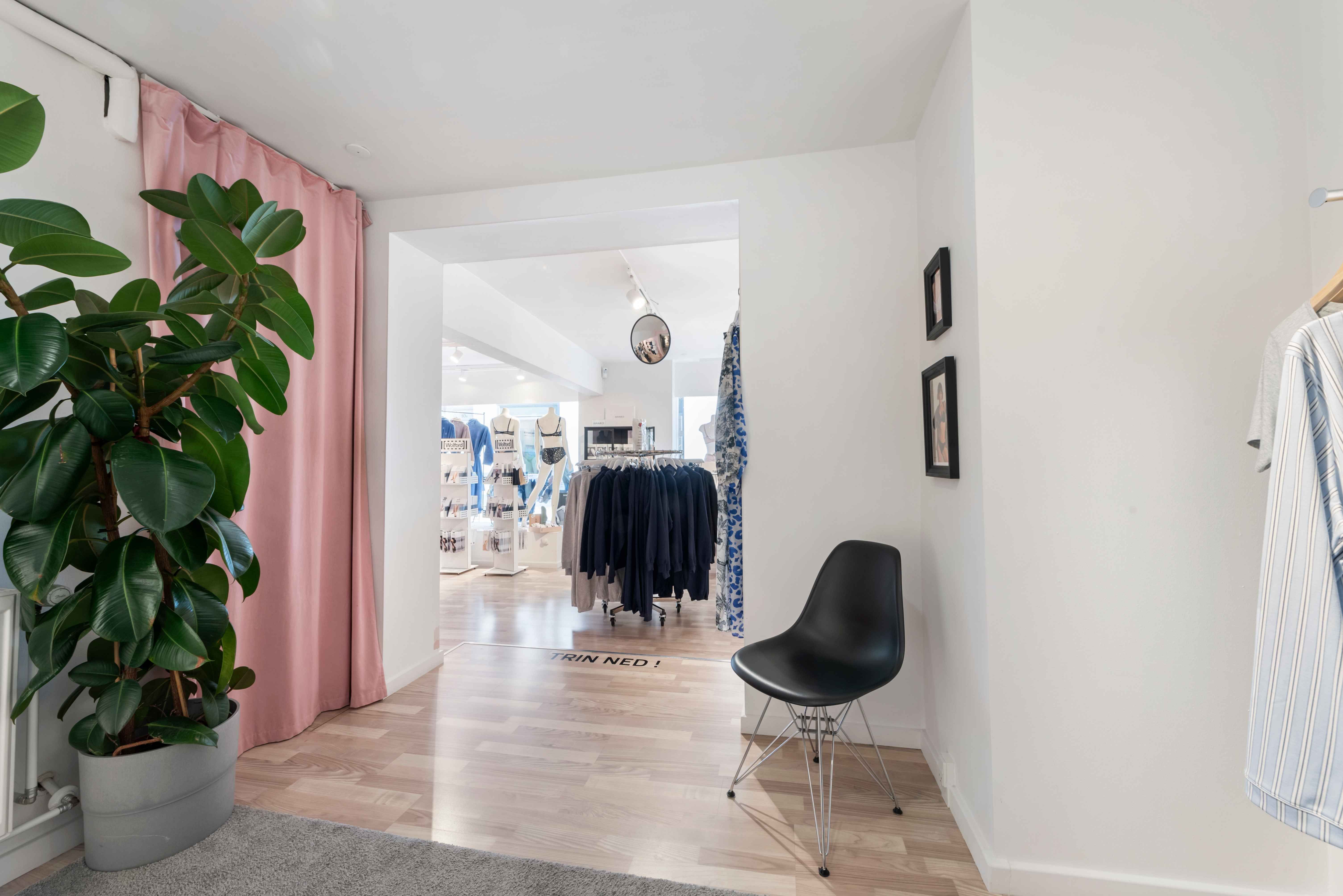 Lys og luftig indgang til butik med trægulv, sort stol og en stor ficus ved siden af. Inde i butikken er der tøj og møbler.