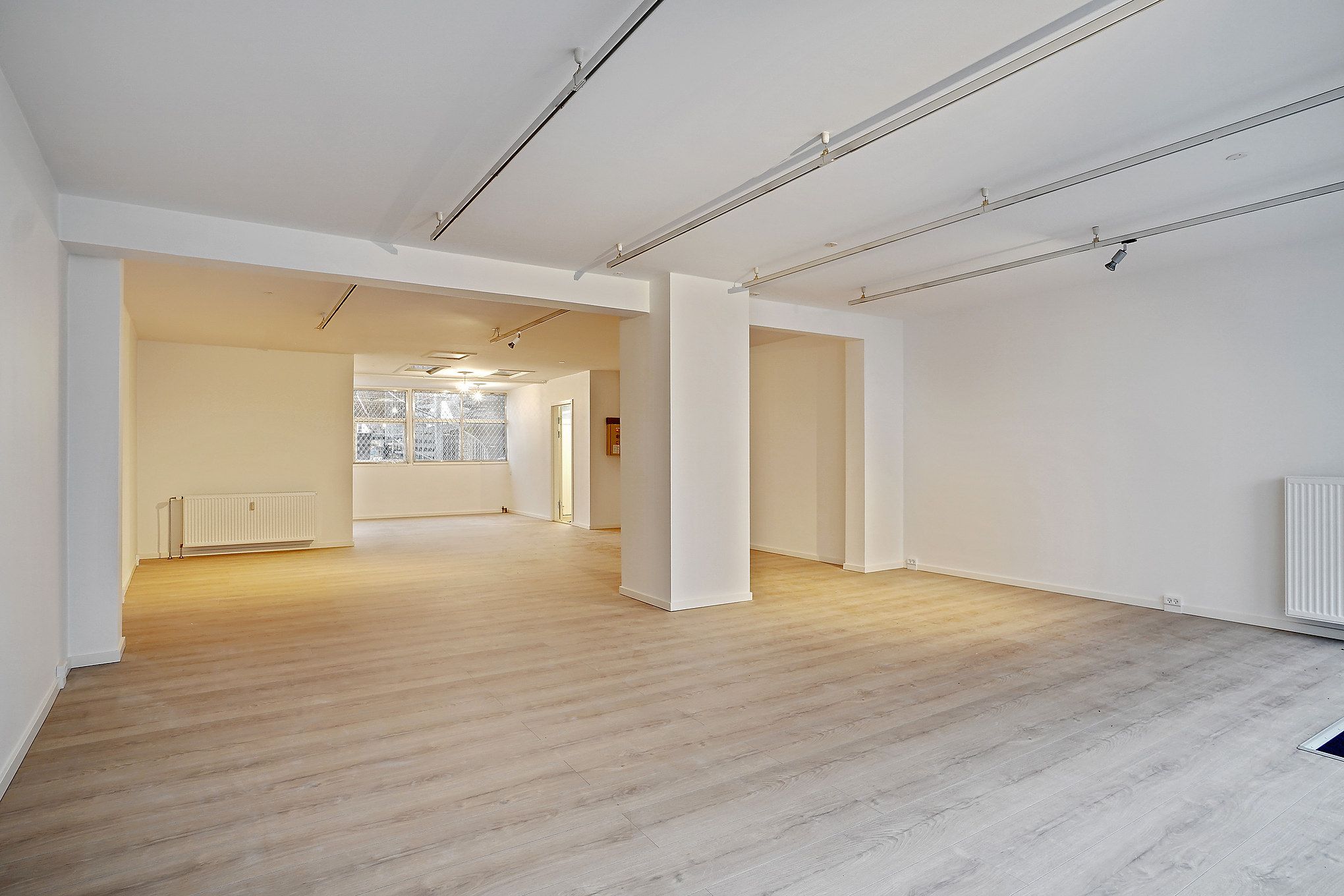 En tom stue med lyse trægulve og hvide vægge. Der er hvide søjler og moderne belysning på loftet. Et vindue giver udsigt til