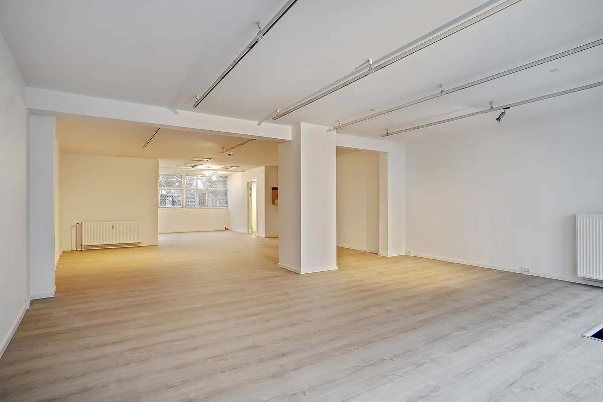 En tom stue med lyse trægulve og hvide vægge. Der er hvide søjler og moderne belysning på loftet. Et vindue giver udsigt til