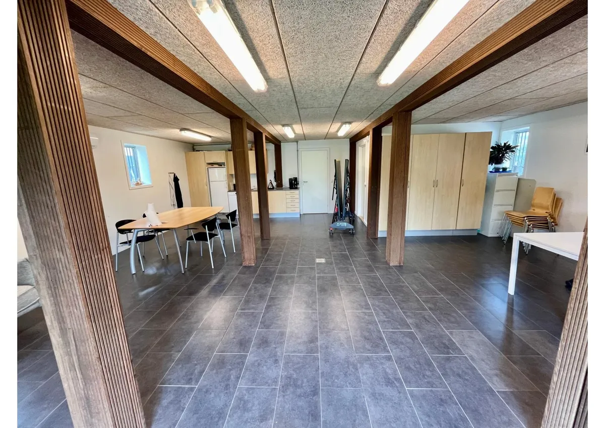 Lyst kontorlokale med træsøjler, moderne møbler, en stor bord, og et køkken med køleskab og kabinetter.