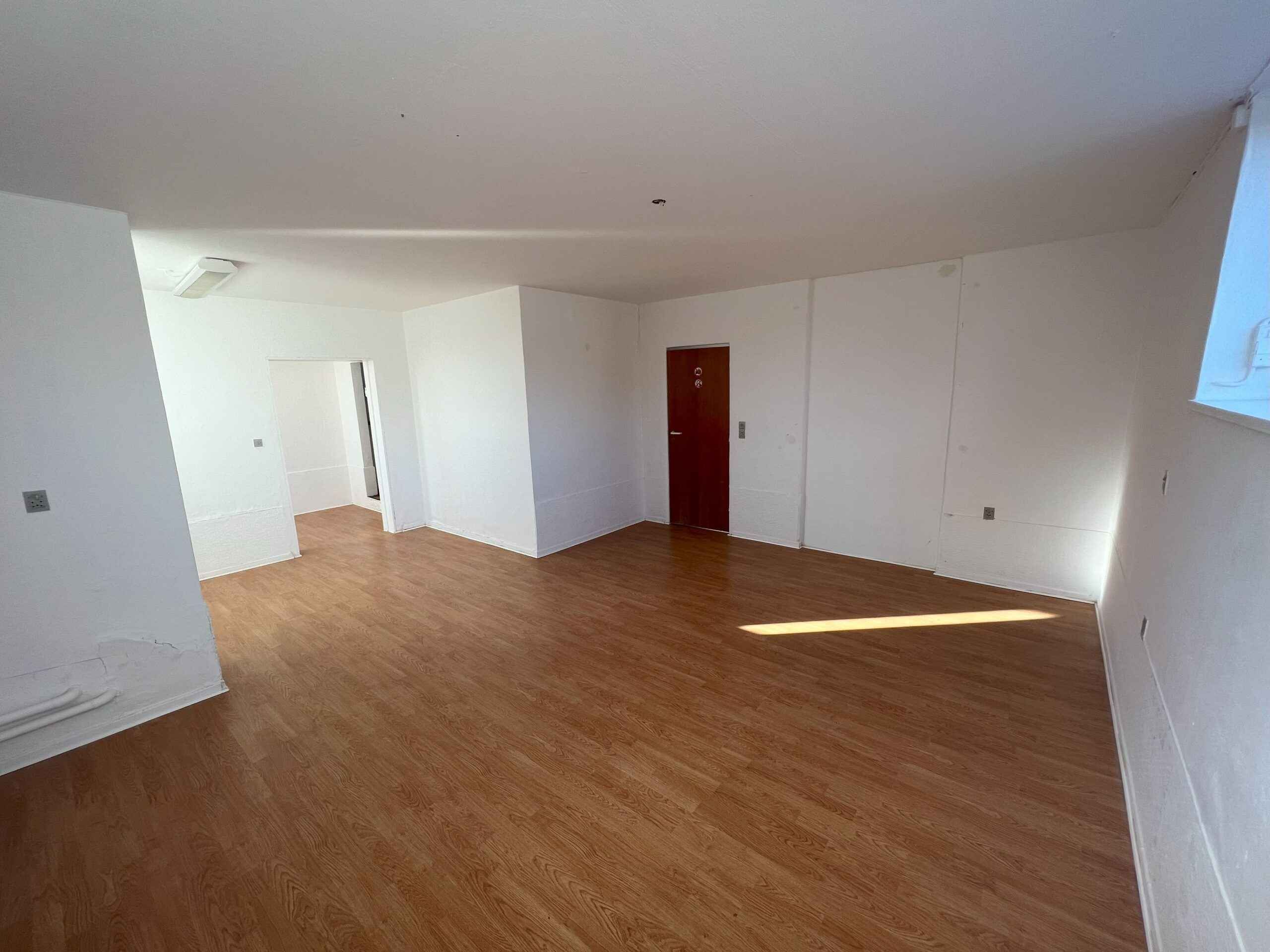 Tomt kontor med lysegult gulv og hvide vægge, samt en rødmalet dør og et vindueskilt til højre.