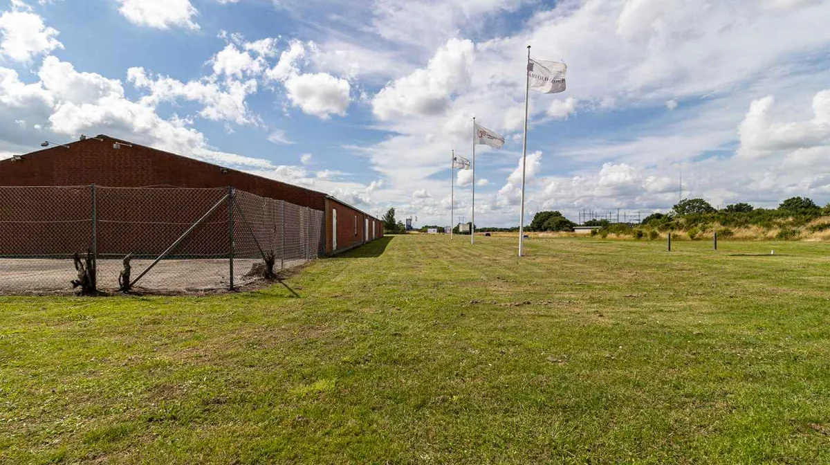 En stor bygning med røde vægge og en lukket port, omgivet af grønne marker og flere flag på flagpæle