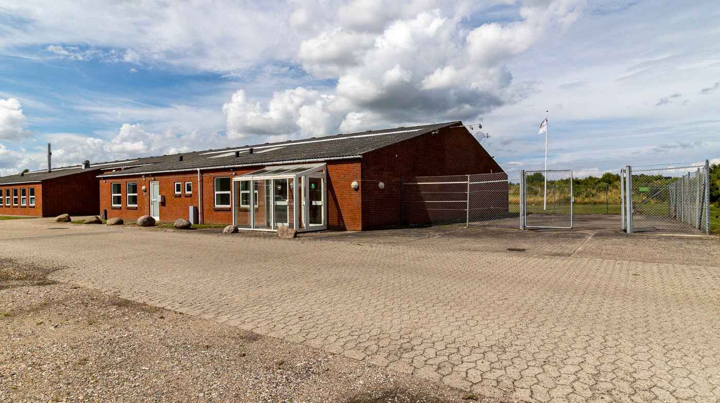 Ledig bygning med murstensfacade og stor glasdør og en stor indkørselsåbning med dobbeltport og flag i baggrunden