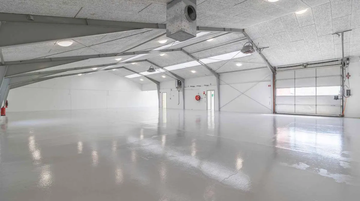 En stor og ren garage med lyse hvide vægge og loft, ventiler og lys, samt en stor hvid garagedør.