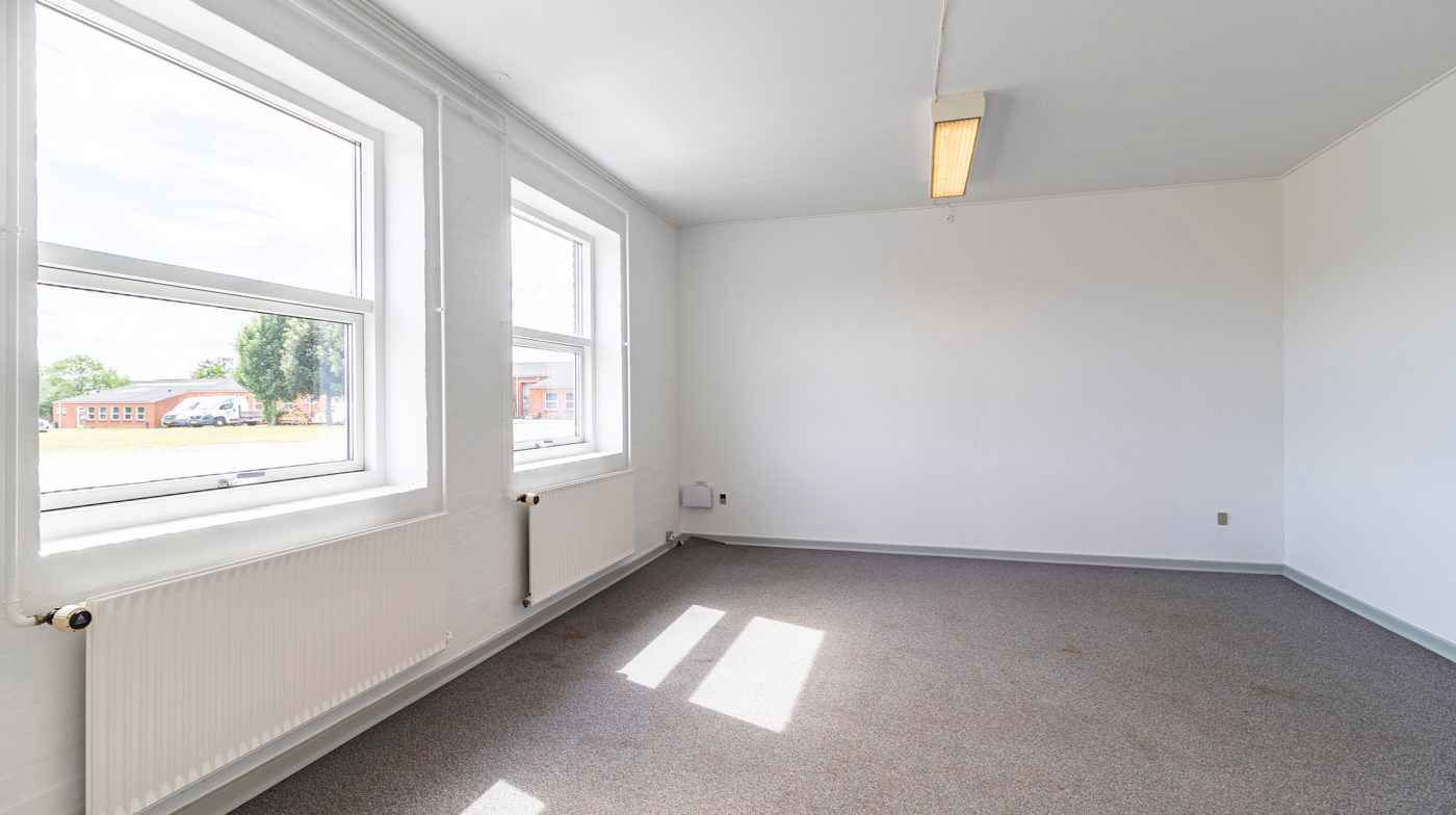 Store lyse kontorfællesskab med hvide vægge, store vinduer og to radiatorer og et gulv med gråt tæppe