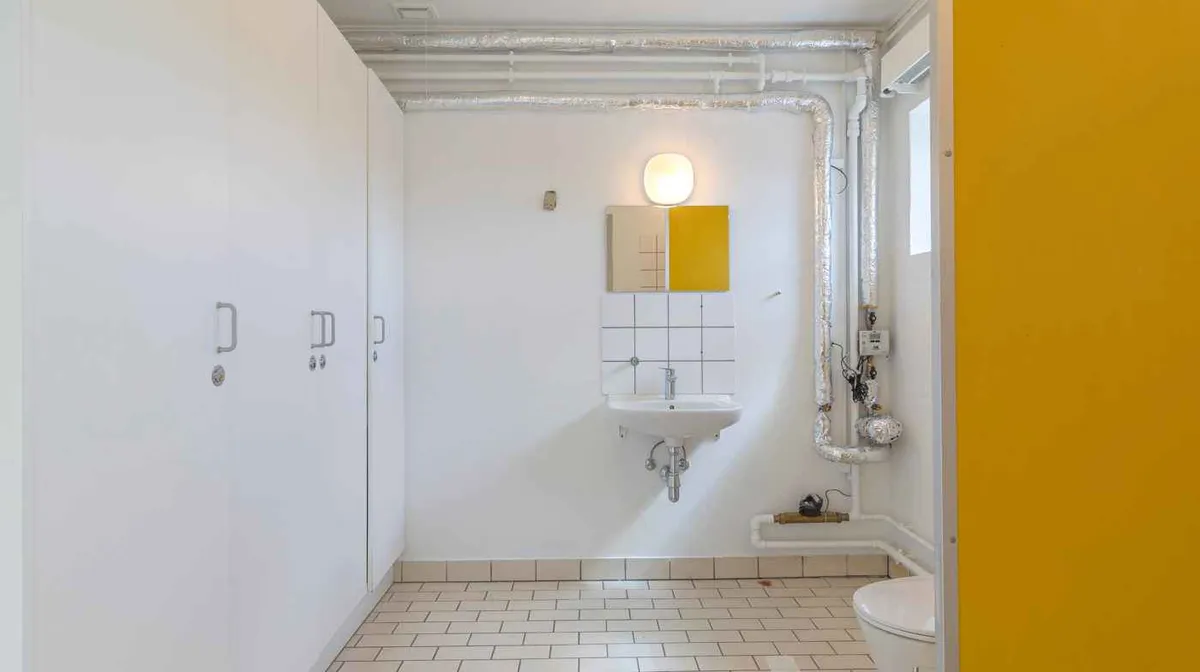 En hvid toiletrum med gult loft og vægge. Rummet har et hvidt toilet, hængende skabe, et hvidt vask og et gult og hvidt