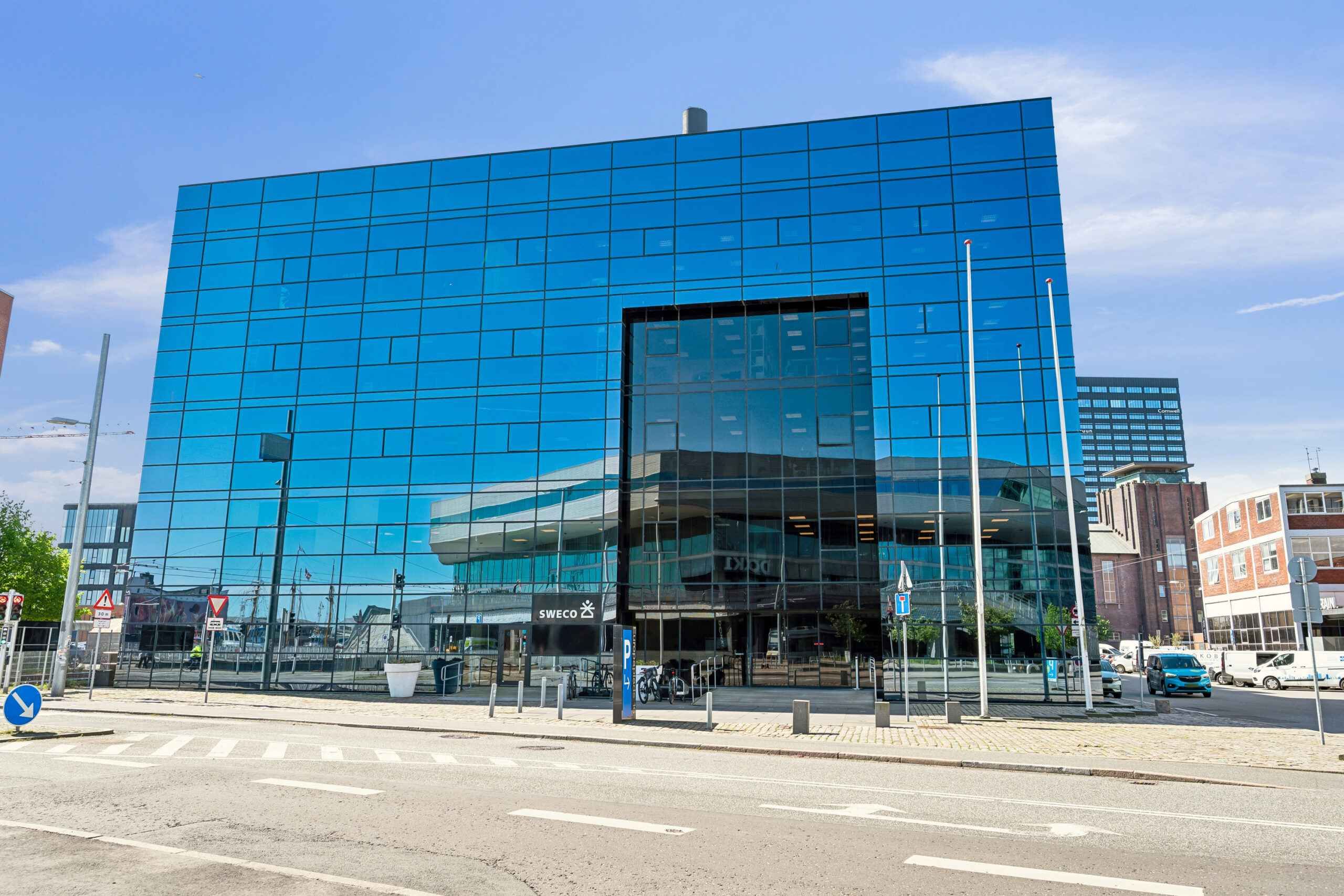 Rummeligt glasbygning reflekterer lyse himmel og skyer; Moderne facade med tydelig tekst og parkerede biler; Bygningens