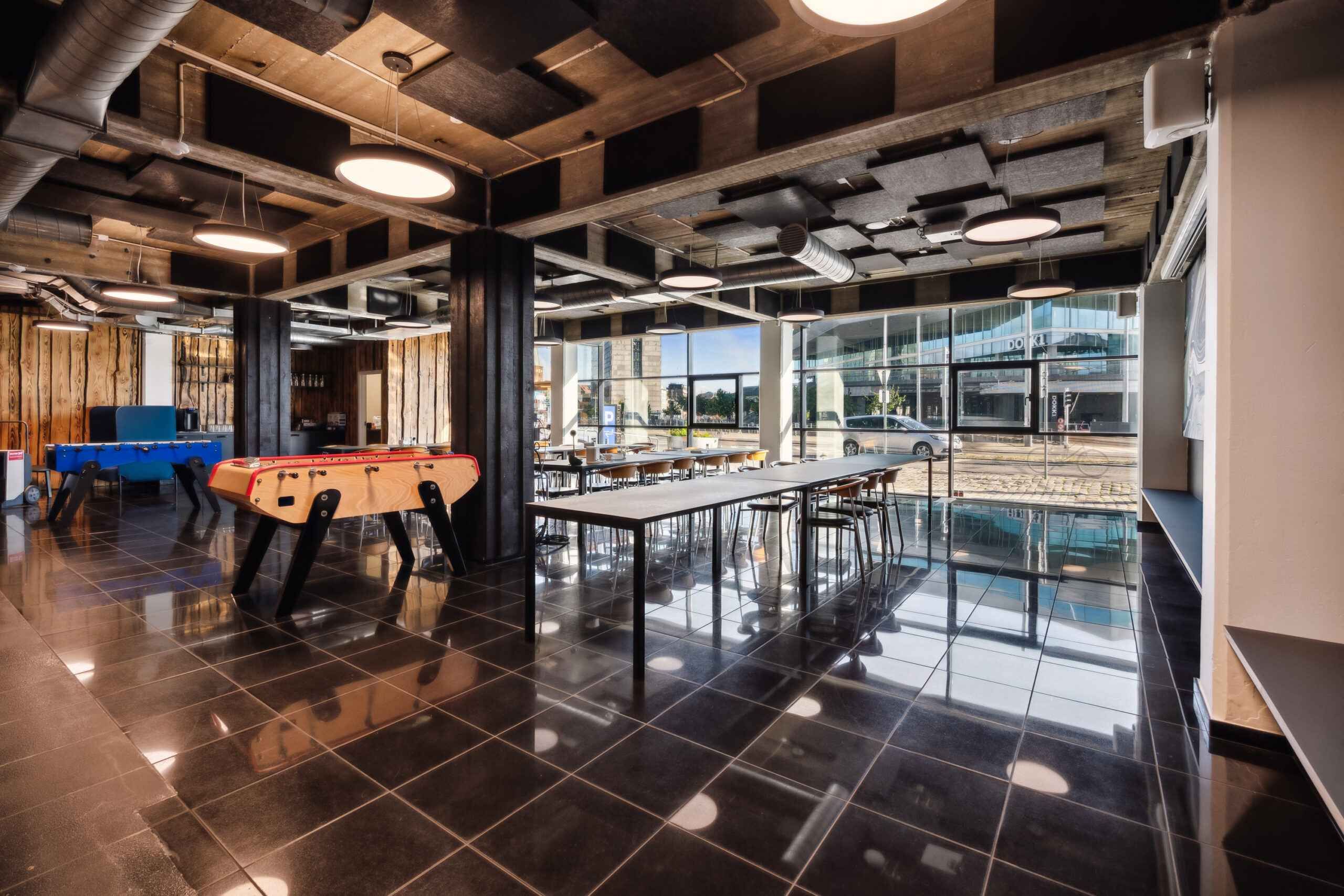 En rekreativ zone med fodboldbord, skrivebord og stole, samt et panoramavindue med udsigt til en parkeringsplads med biler