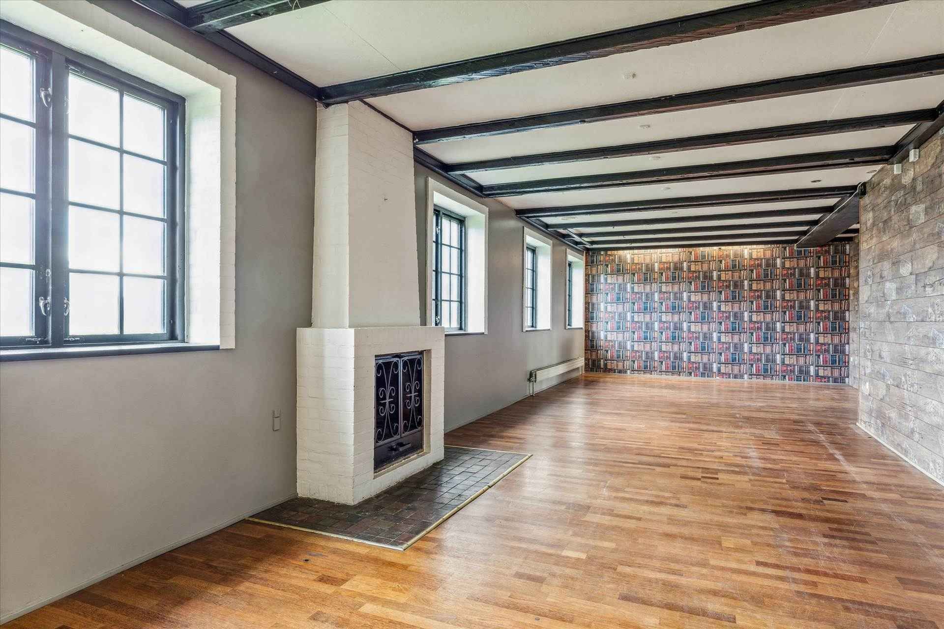 Et stort rum med lyse trægulve og højt til loftet. Et hvidt, moderne skab med et indbyggede ildsted. Et biblioteksvæg med