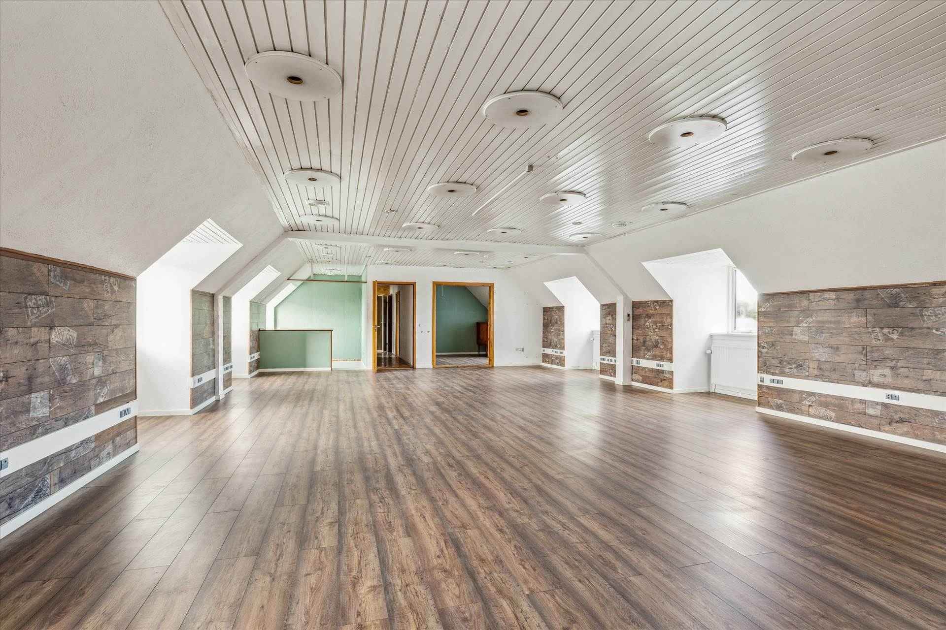 En stor værelse med trægulv og loft med hvide loftbjælker, flere runde loftslamper, og træbeklædte vægge i enden af rummet