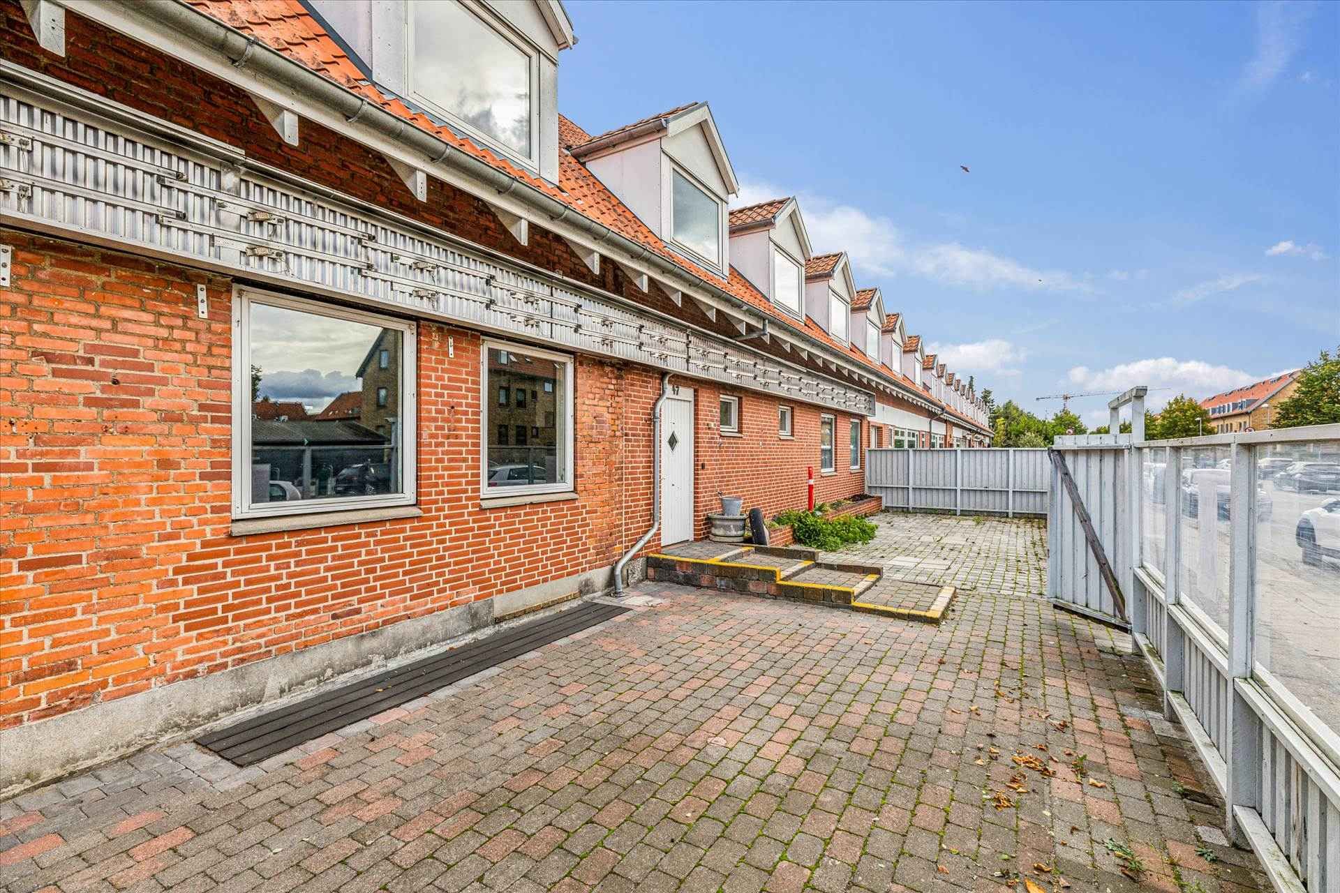 Murstenshus med hvide vindueskarme og glasruder, åben dør, skrappe trin, og en lille grøn plante ved siden af.