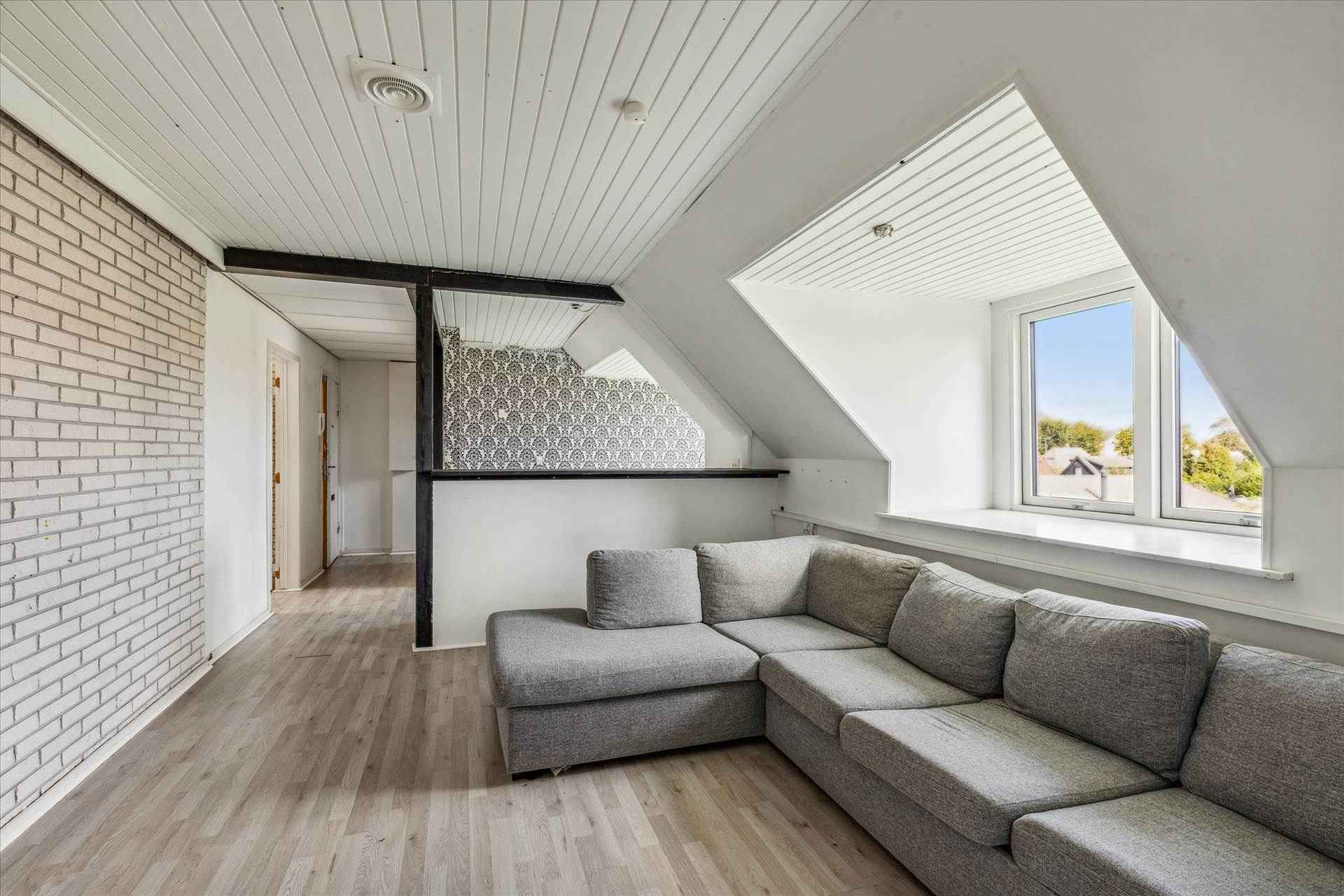 Et enkeltværelse med hvidt loft og gulv og en L-formet sofa med tre puder foran et vindue