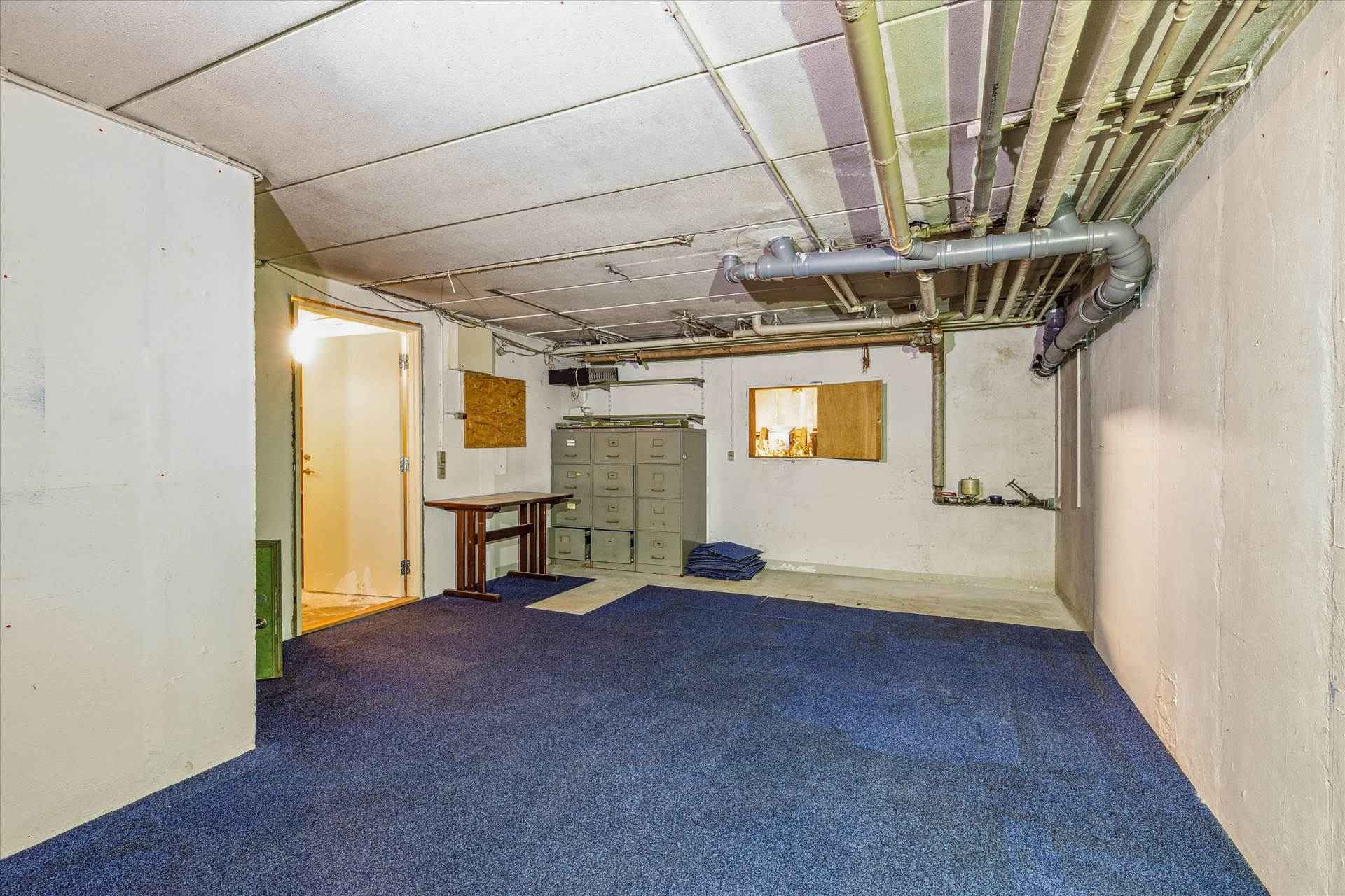 Et stort, lyserødt gulvtæppe dækker gulvet i et rum med hvide vægge og lysegrå loft. Et træbord og en skabgruppe er placeret