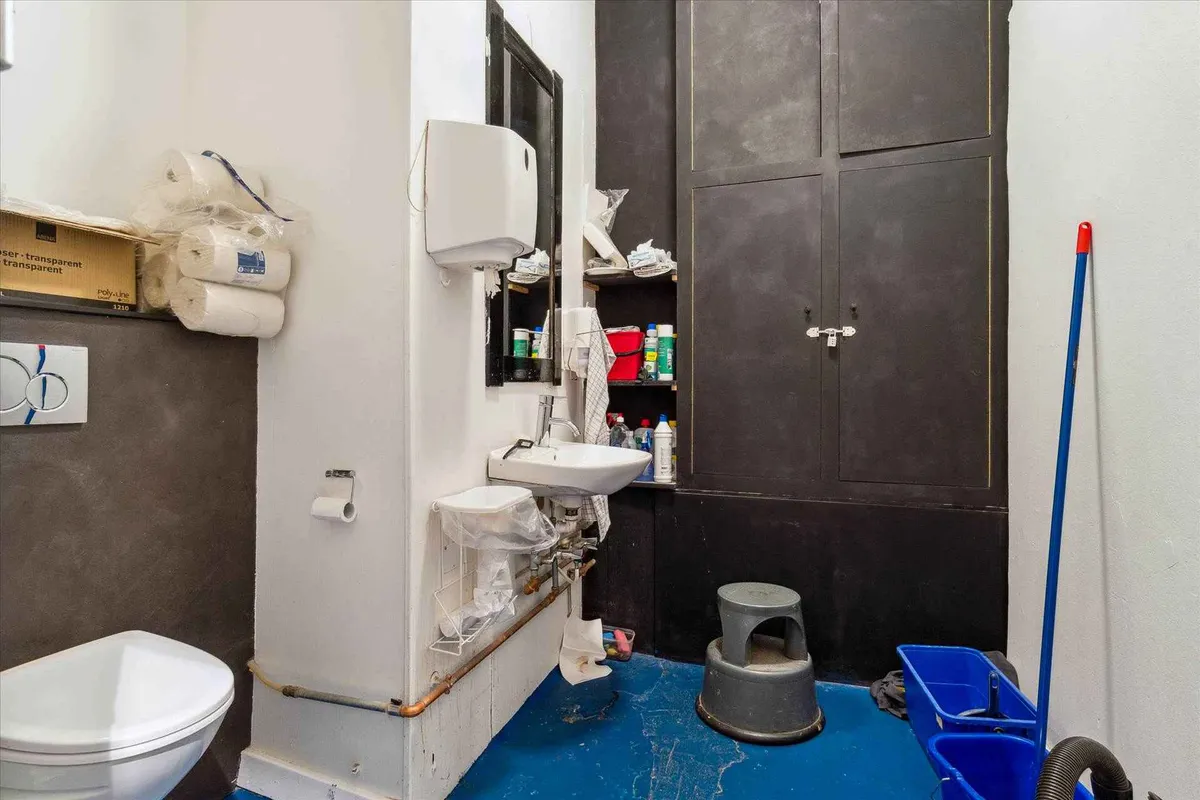 Rum med toilet, håndvask og rengøringsudstyr. Toilet med papkasser på toiletbordet. Håndvask med to hætter og en