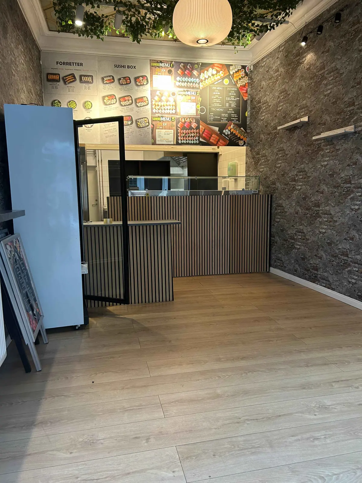 Sushibar med trægulv og glasvæg. Der er et køleskab, menukort og lysekroner. Der er et bord med to lyse lysestager