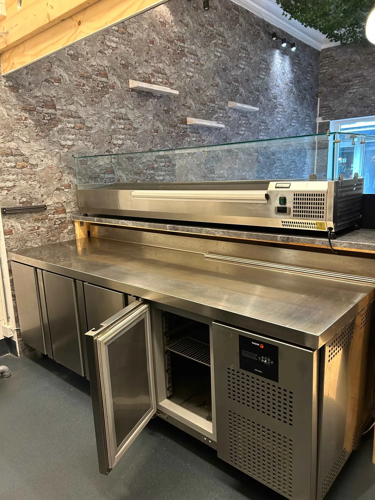 Professionel køle- og frysekombination med glasfront og indbygget opvarmer, placeret i kommercielt køkken med røde mursten