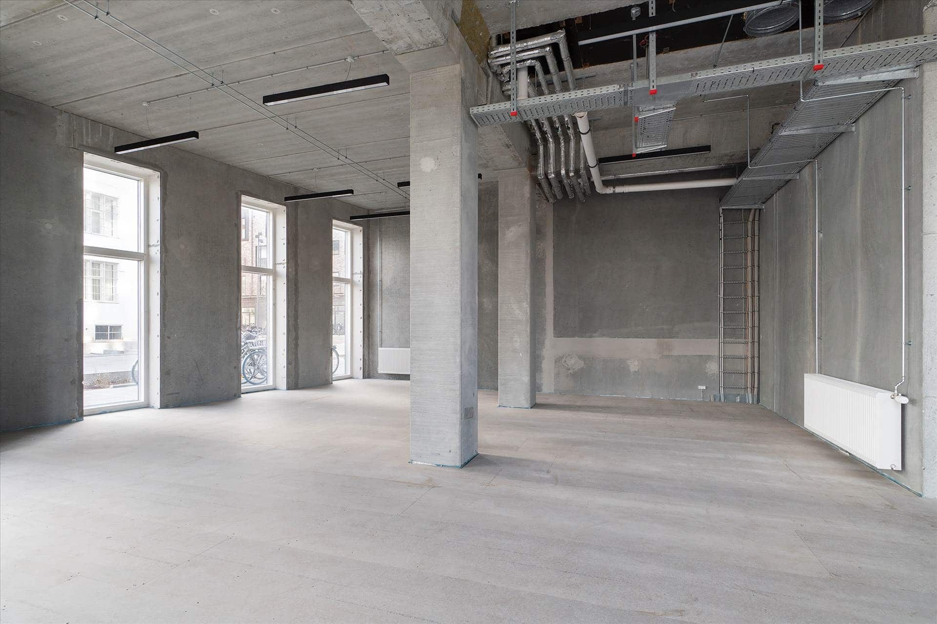 En stor, tom erhvervslokal, med store vinduer og høje lofter, samt en række rør og ledninger på loftet
