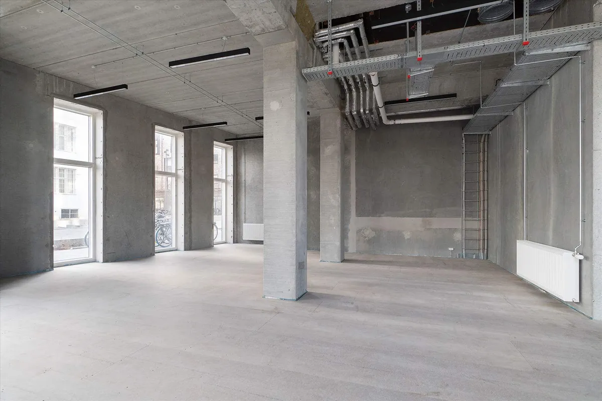 En stor, tom erhvervslokal, med store vinduer og høje lofter, samt en række rør og ledninger på loftet