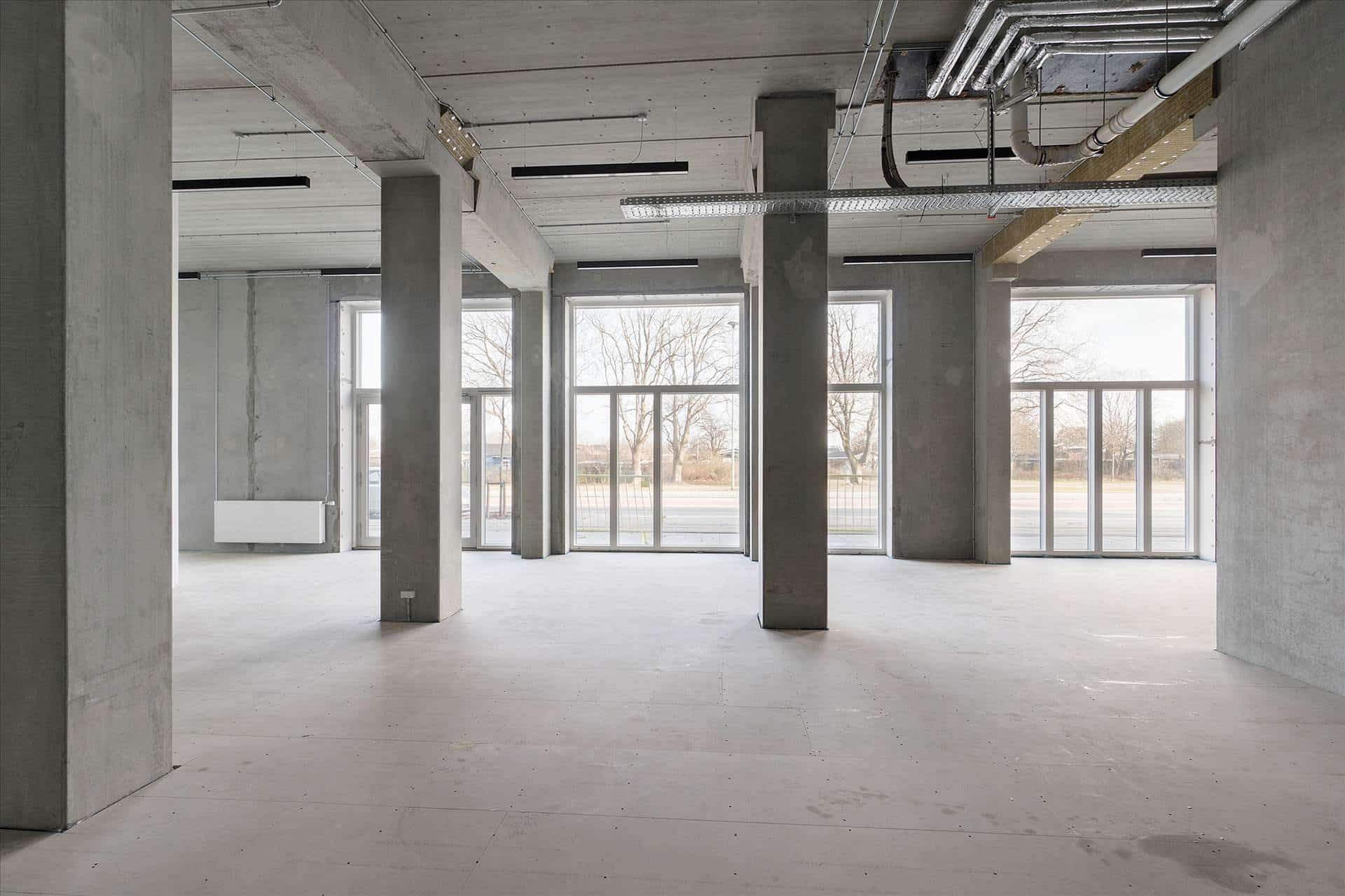 Store kontorlokaler med højloft og glasvinduer mod udendørsområde med parkeringsplads og grønne træer i baggrunden