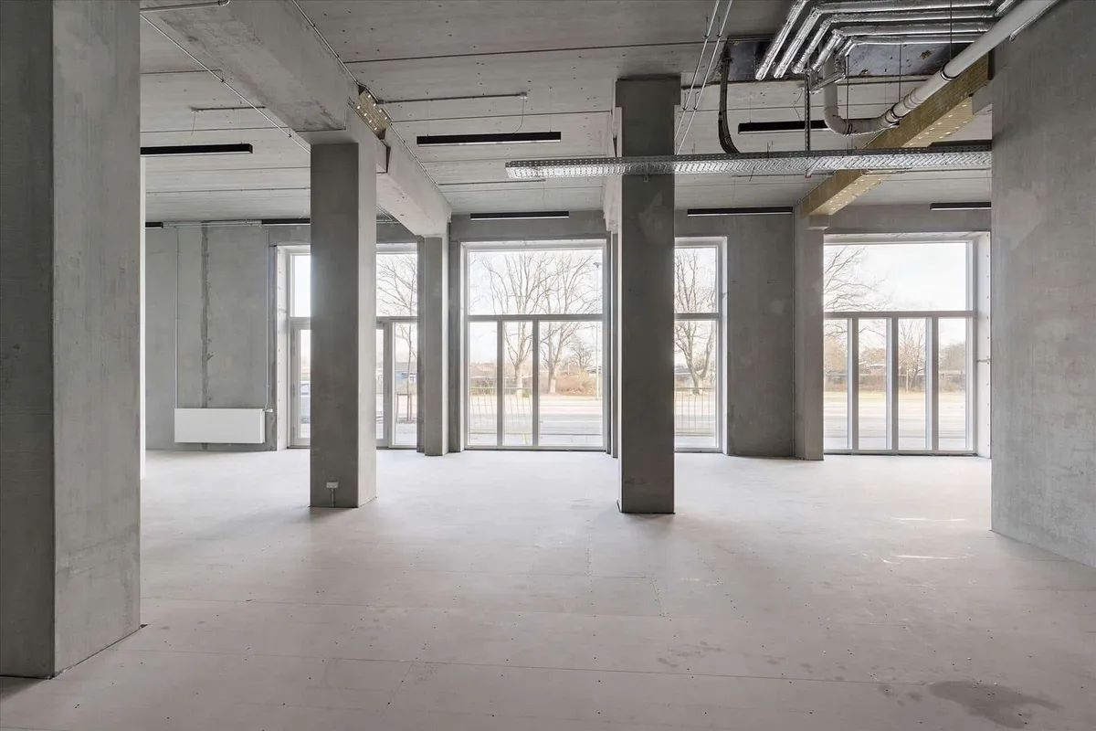 Store kontorlokaler med højloft og glasvinduer mod udendørsområde med parkeringsplads og grønne træer i baggrunden