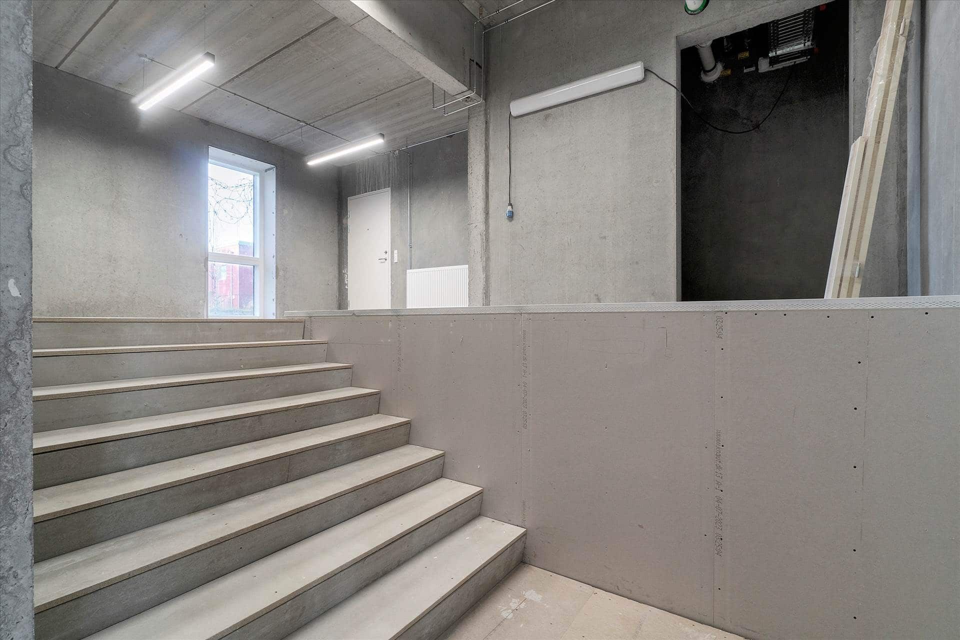 Tre stegede trapper af beton i et tomt lokale med en åben dør og et vindue samt lys i loftet og en hvid mur i baggrunden