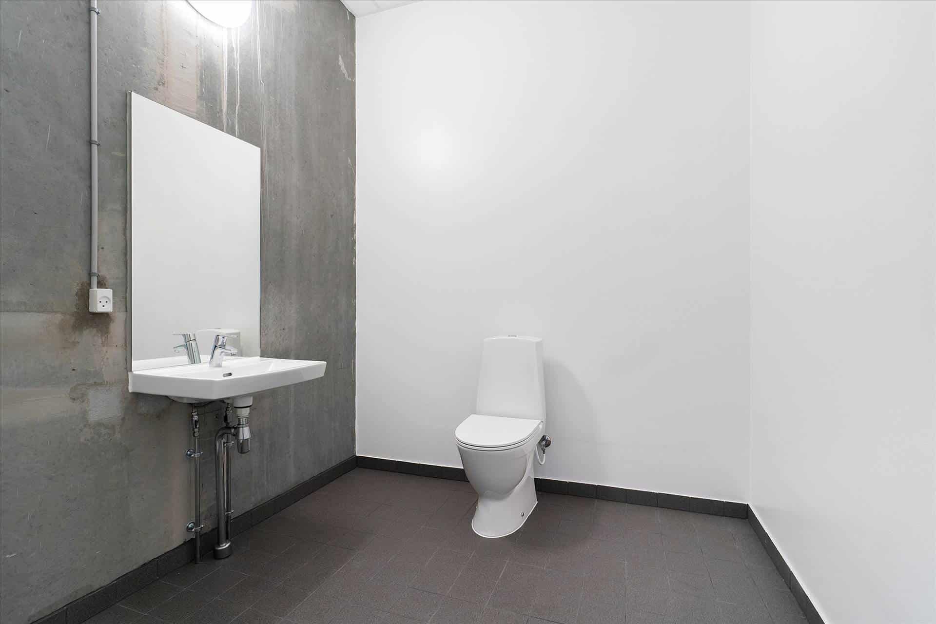 Toiletterum med hvidt toilet og hvid vask med 2 vandhaner, og en spejl, hvor der er en stikkontakt på væggen