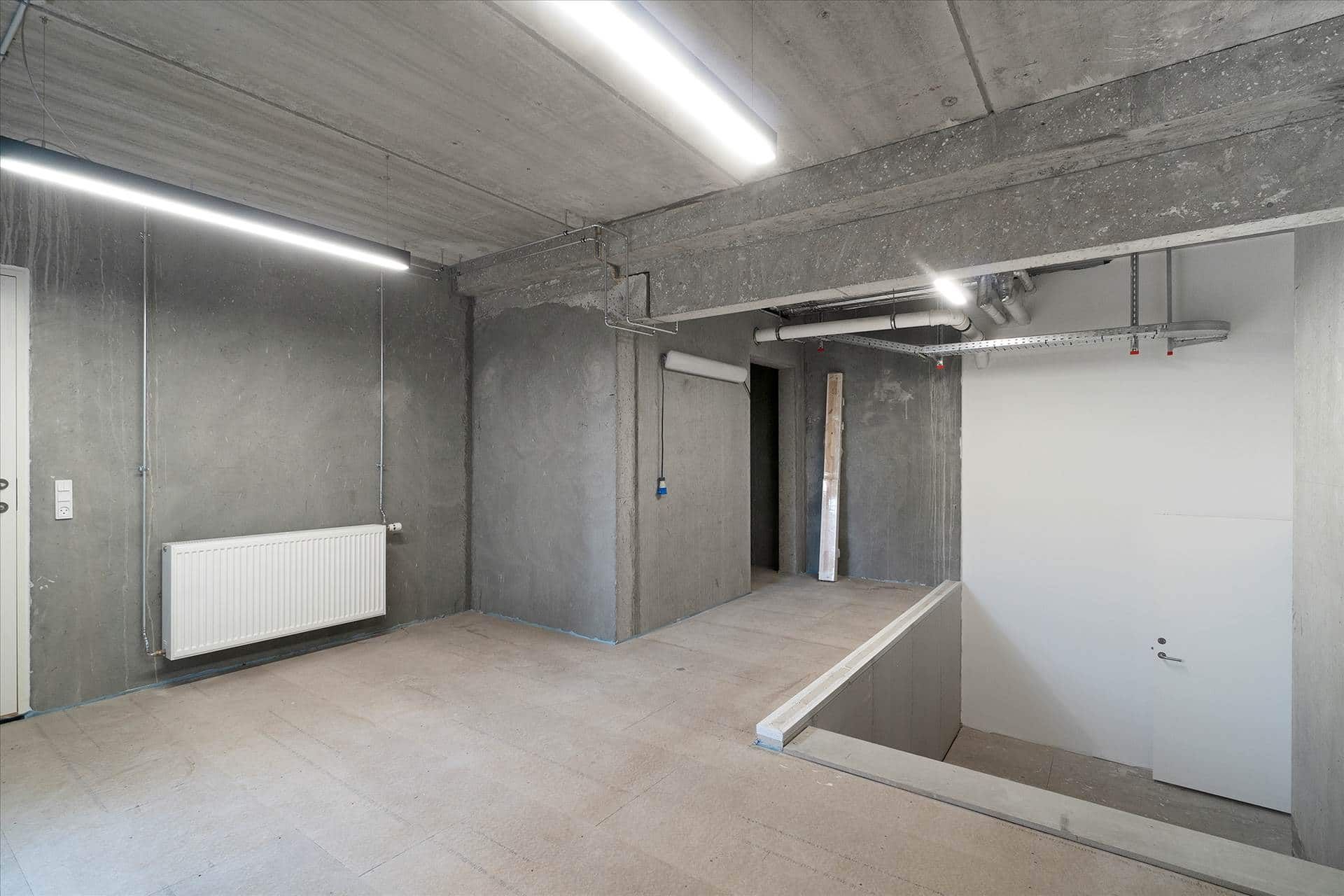 Rum med lysegul gulvbelægning og hvide vægge. Der er en radiator og to lysestager på loftet. Der er en åbning til en trappe