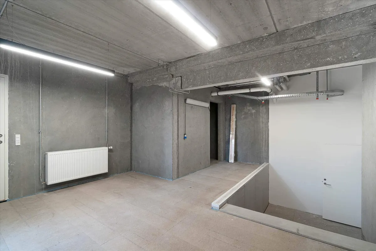 Rum med lysegul gulvbelægning og hvide vægge. Der er en radiator og to lysestager på loftet. Der er en åbning til en trappe