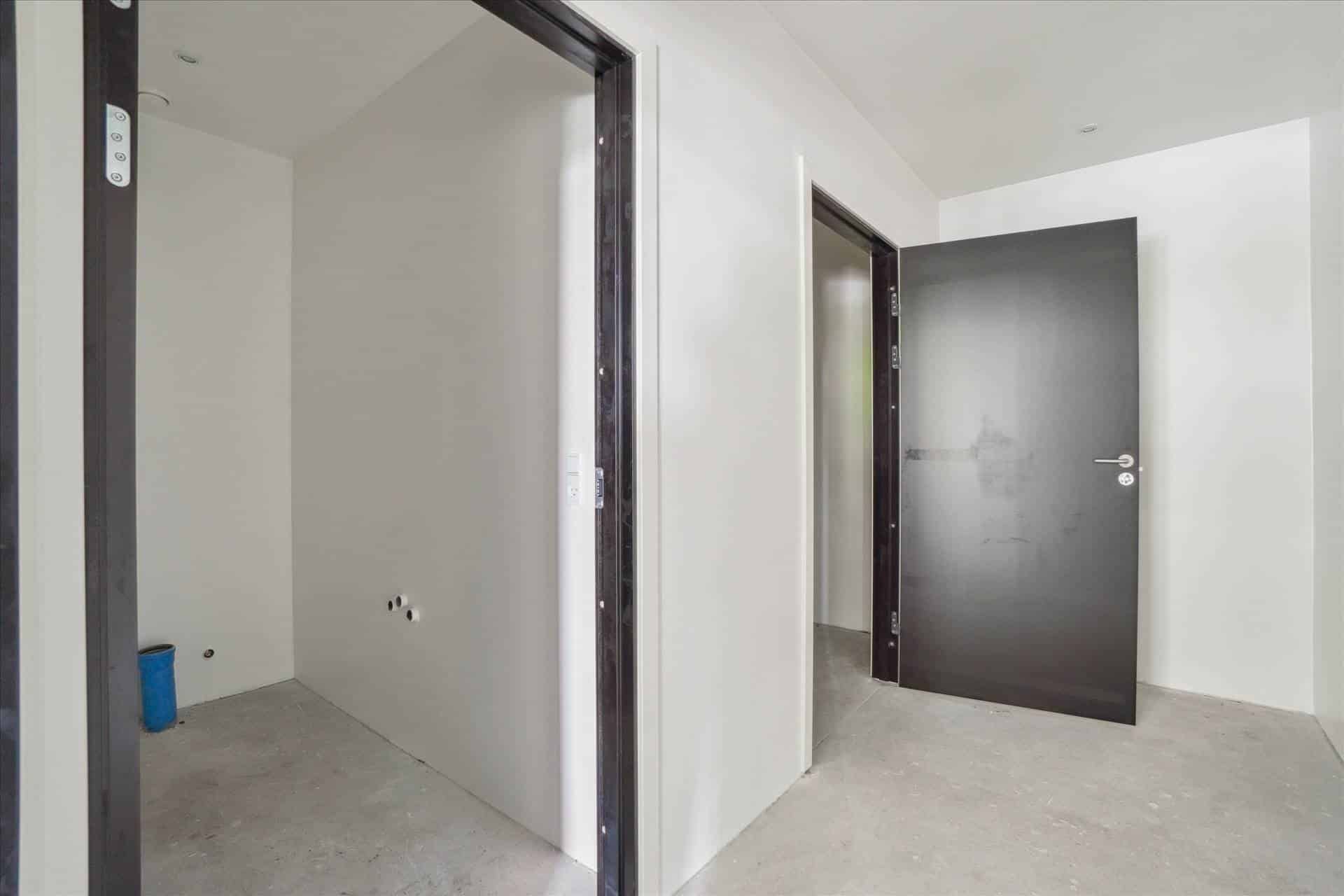 To store, hvide vægge og en åben dør. En lille, blå søjle står i hjørnet. Gulvet er ujævnt og ufuldstændigt.