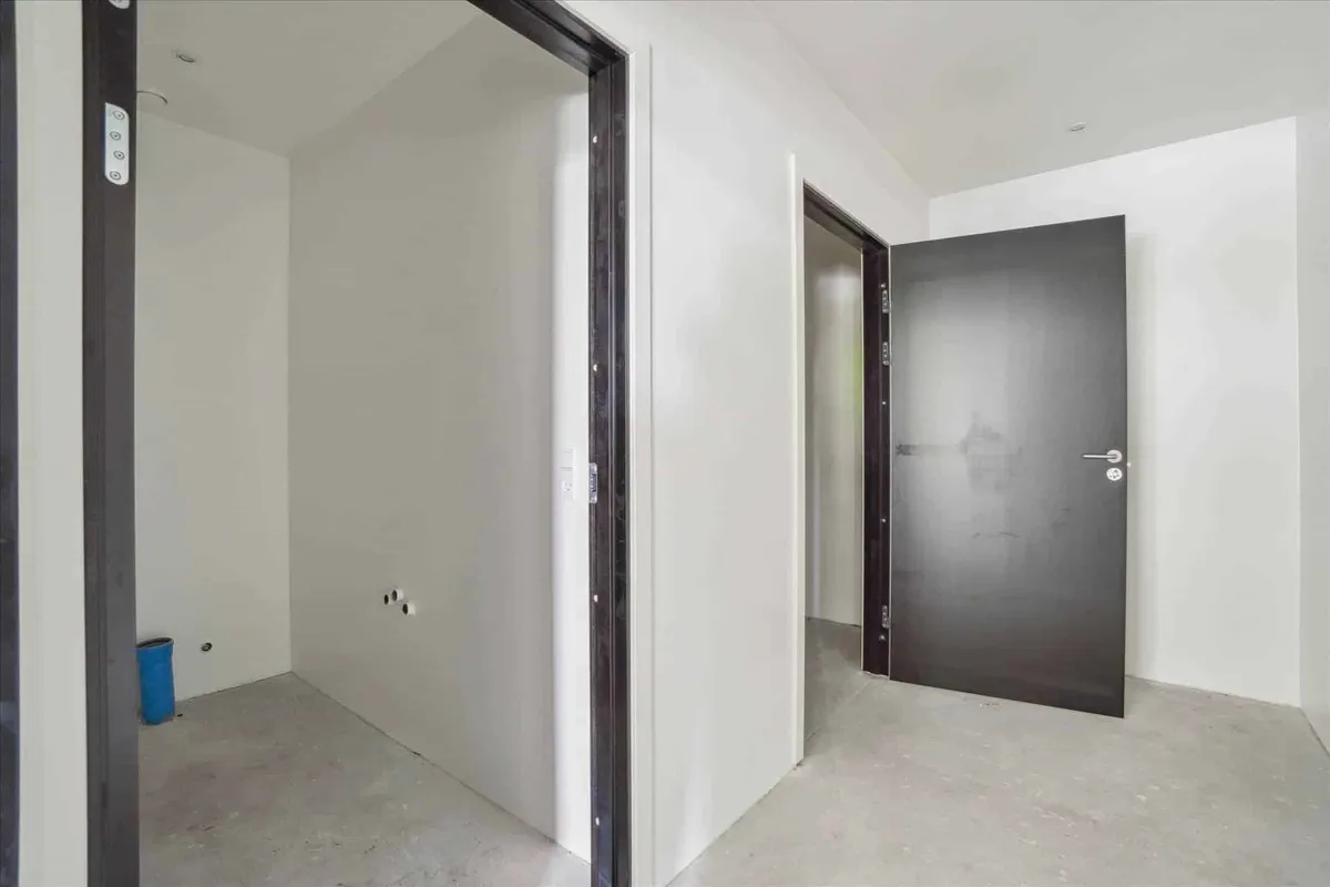 To store, hvide vægge og en åben dør. En lille, blå søjle står i hjørnet. Gulvet er ujævnt og ufuldstændigt.