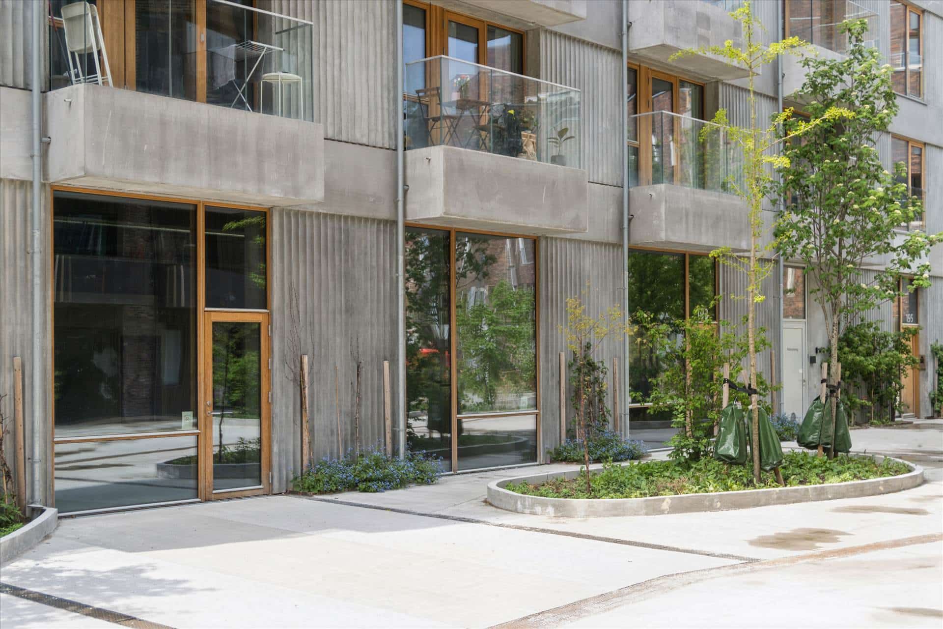 Lejlighedsbygning med store glaspartier og terrasser. Glasskærme og trævinduer. Fortove med grønne plantager. Balkoner med