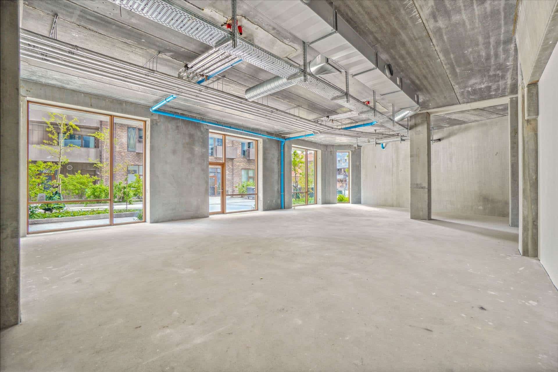 Lejemål med store vinduer og højloft i København. Betongulv og ventiler i loftet. Udsigt over grønne områder og bygninger